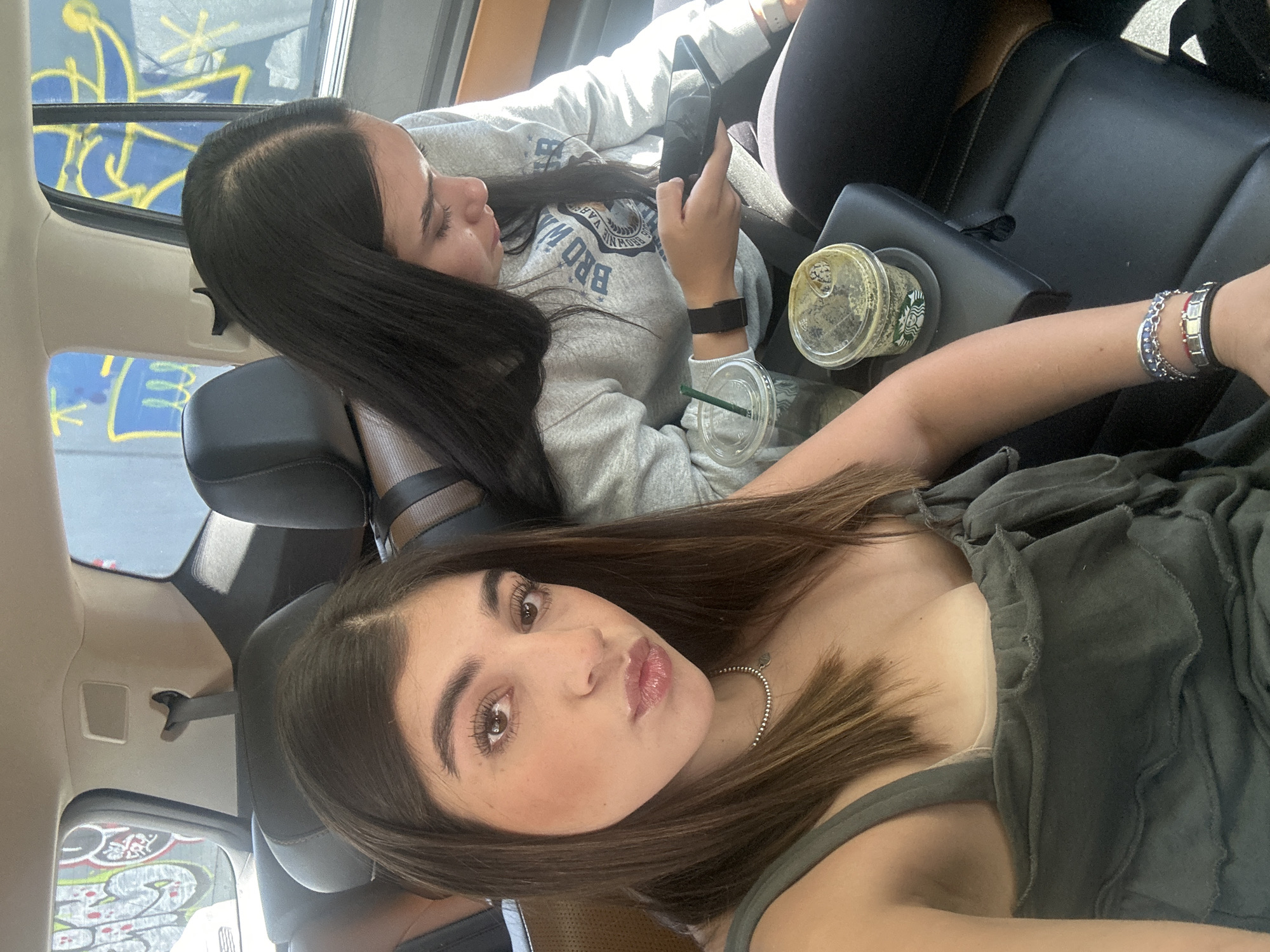 The image shows two young women in a car, likely taken in Mexico City given the graffiti visible through the window, a common feature in urban areas of Mexico. The woman closer to the camera is wearing a dark green ruffled top and a silver necklace, and she is looking towards the camera with a slight pout. Her hair is shoulder length, and brown with light highlights. The other woman is in the back, wearing a grey sweatshirt with "BROWN UNIVERSITY" printed on it, and black leggings. She is holding a phone in her hands. Between them is a drink from Starbucks. Both seem to be relaxed. The car has black leather seats. The lighting is bright, possibly midday.