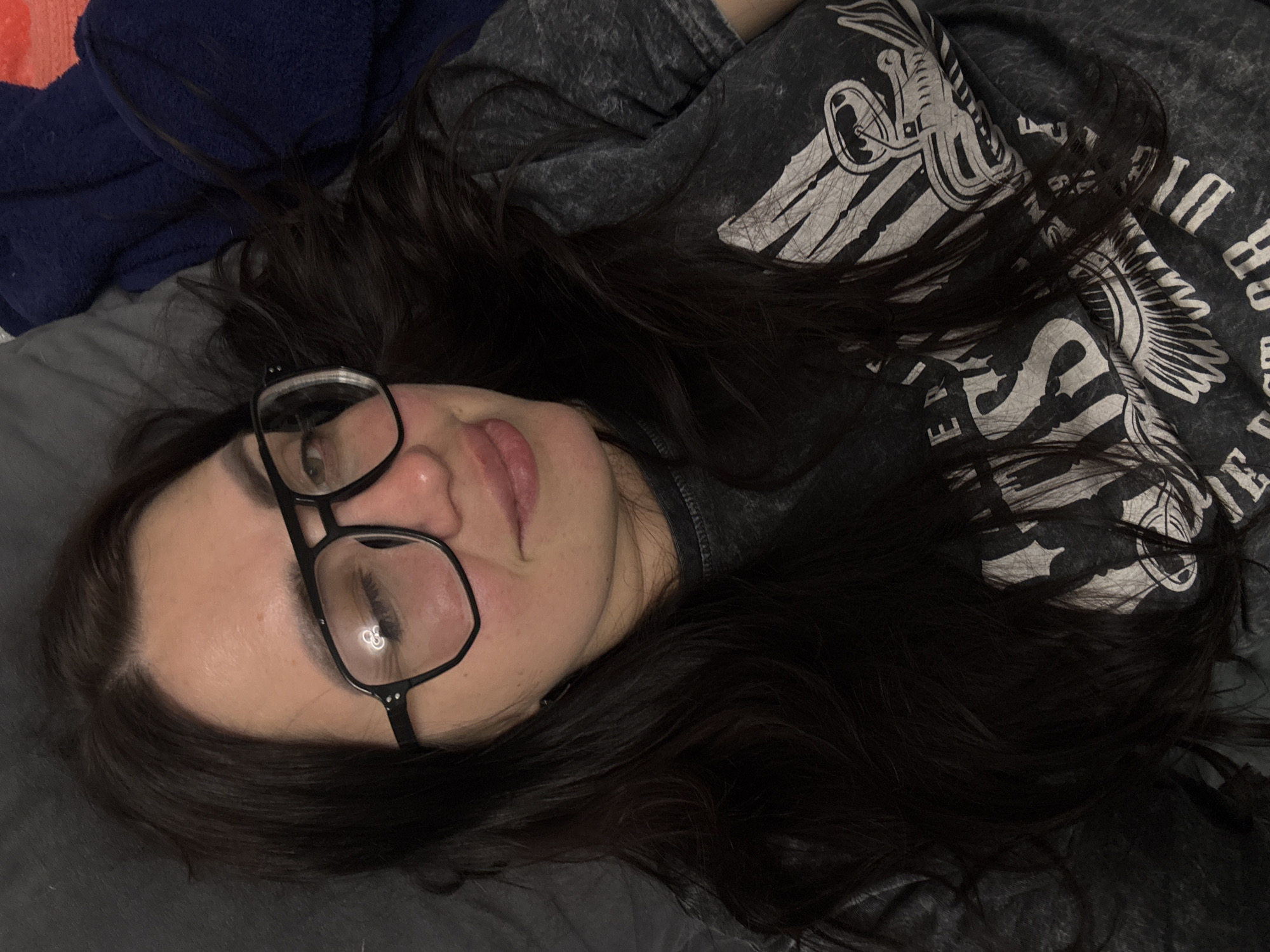 In the city of Unknown, Unknown, an overhead shot captures a single female individual in a reclined position. The subject, with long, dark brown hair, wears large black-framed glasses; a circular light reflection is visible in the left lens. Her right eye is closed in a wink, while her left eye is open, revealing a light-colored iris. A slight smile is present, with her lips slightly parted. She is wearing a dark grey, possibly acid-washed, t-shirt featuring a large white graphic print that includes partial text such as "ERS", "S", "197", and "OR", along with stylized imagery resembling wings or an eagle. The background to the left consists of a plain grey fabric, likely a pillow or bedding. To the right, a dark blue textured fabric, possibly a blanket, is visible, with a small portion of an orange-red fabric underneath it.