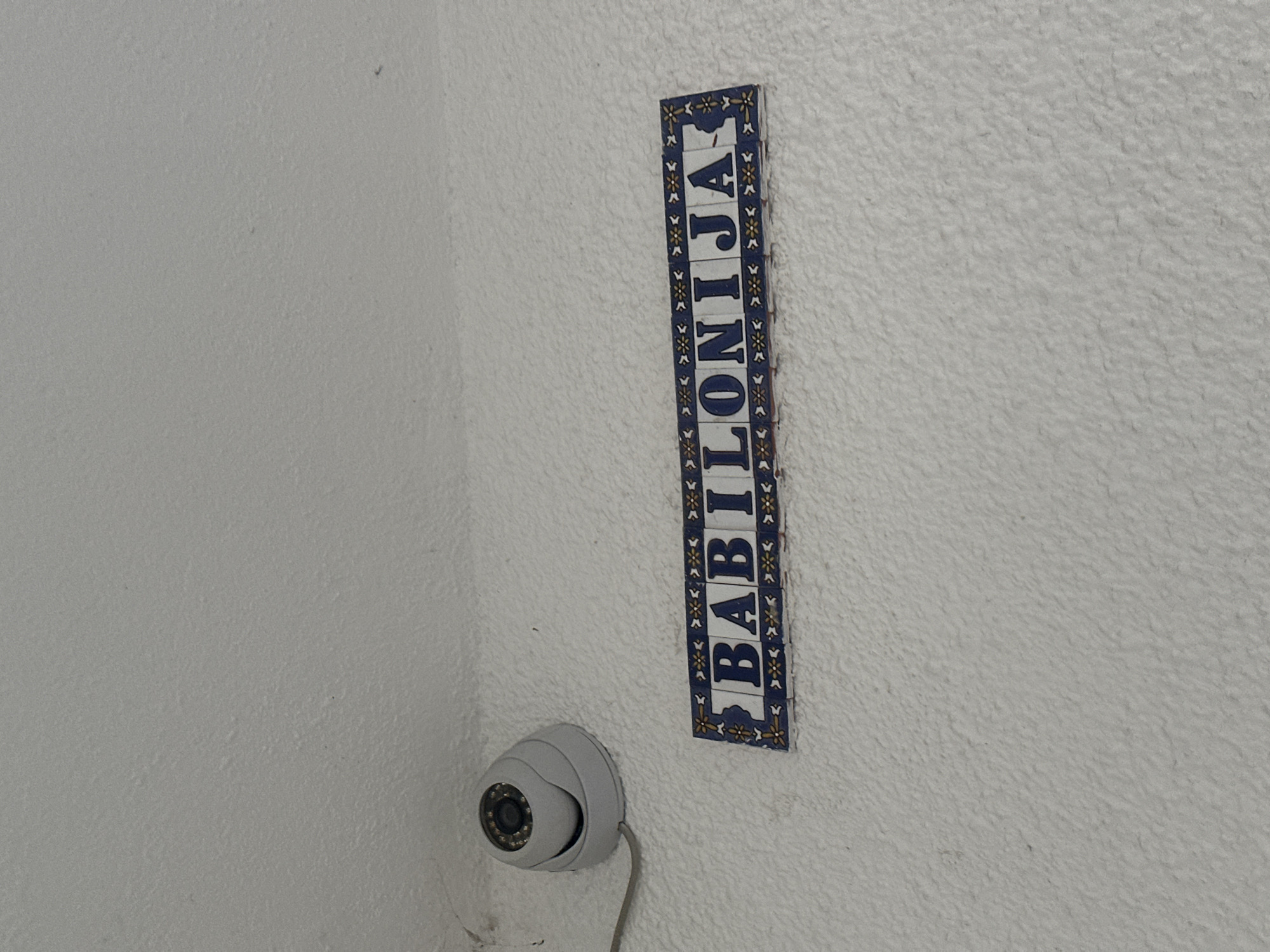 A white, textured wall features a vertical, tiled sign reading "BABILONIJA" in blue letters. To the left and below the sign is a white dome-shaped security camera with a visible lens and a gray cable extending downward. The location is likely Budva, Montenegro, as "Babilonija" is a street or place name there.
