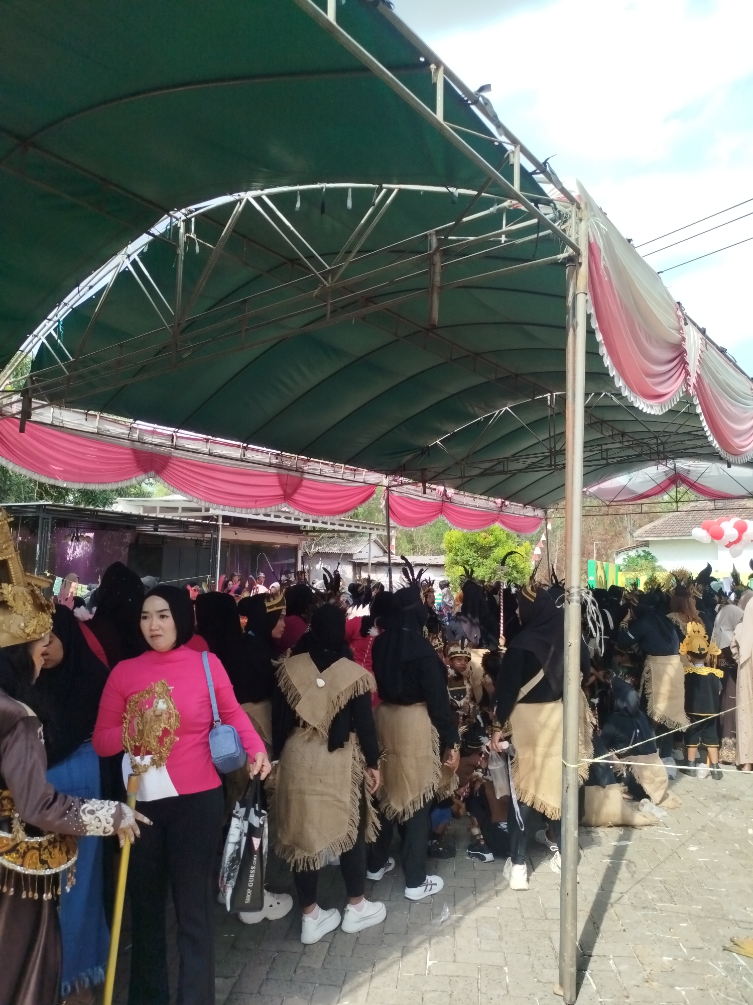 In Jambu, Indonesia, under a large green tent with pink and white drapes, a crowd gathers for an event. Several people are dressed in traditional attire, including some wearing black headscarves and burlap-like skirts with fringe. One woman in the foreground wears a bright pink long-sleeved shirt and black pants, holding a decorative golden object and a blue bag. Another person to her left is dressed in a brown costume with gold embellishments. In the background, a stage area is visible with more people in costumes and a white structure. Red and white balloons are visible in the upper right corner of the image, suggesting a festive occasion. The ground is paved with light-colored bricks.