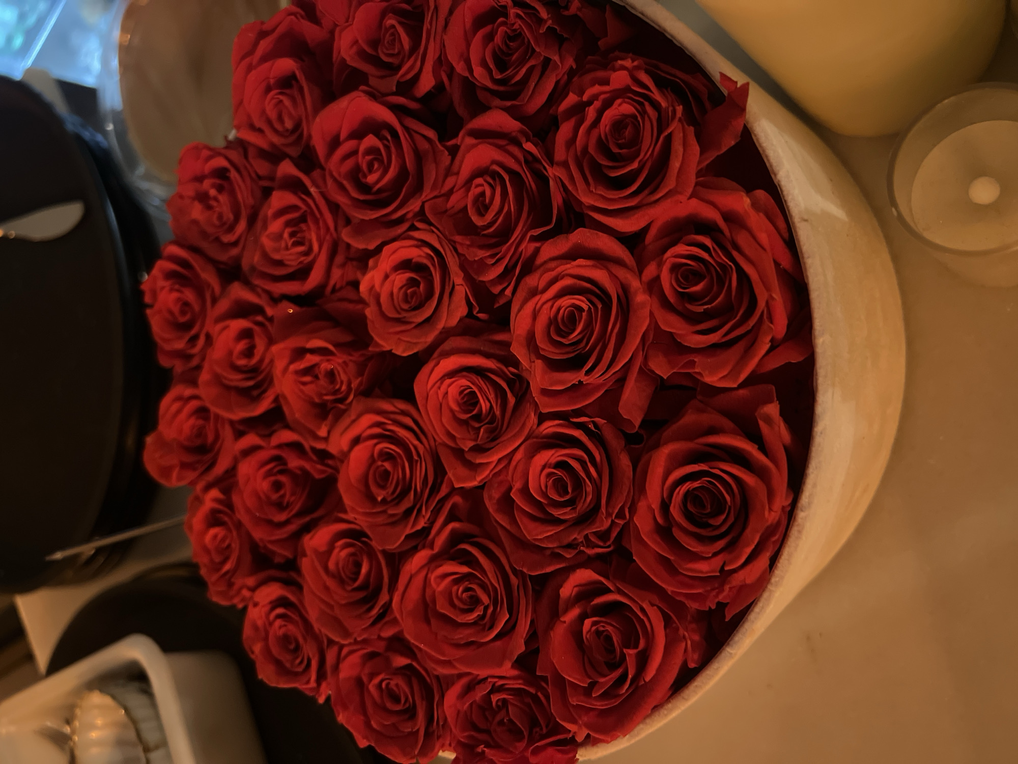 An overhead photograph captures a lush arrangement of deep red roses displayed in a light-colored, possibly cream or beige, round container. The roses, tightly packed and uniform in appearance, feature rich, velvety petals, suggesting they may be artificial or preserved.
To the left of the rose arrangement, a dark, reflective surface, likely a table or tray, is visible, with a metallic utensil handle glinting subtly in the soft light. The right side of the frame reveals several warm-toned, possibly ceramic or wax, decorative objects, including a cylindrical item and a small, clear glass votive candle holder containing a lit candle.
The overall lighting is soft, warm, and ambient, casting a gentle glow that suggests an intimate evening or nighttime setting. The scene evokes feelings of romance, celebration, or comfort, implying a special occasion or a decorative display. No text is visible in the image. The setting is possibly in Barcelona, Spain.