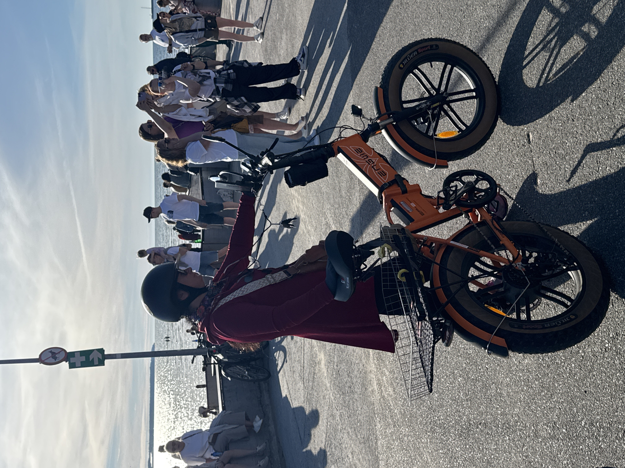 The image depicts a sunny day along the coastline in Tallinn, Estonia, with a focus on an electric bicycle and its rider. The person, wearing a helmet and a maroon jacket, stands next to an orange fat-tire electric bicycle. The bicycle has a front basket and appears to be parked on a paved surface, possibly a promenade, with shadows cast by the sun.
In the background, a body of water, likely the Baltic Sea, shimmers under the bright sky. Several people are visible, some sitting on a low wall, others standing and interacting. A street sign with a dog prohibition symbol and an arrow is positioned on a pole to the left of the frame. The overall atmosphere suggests a leisurely day out in a public space by the sea. The location is identified as Tallinn, Estonia, based on the context provided.