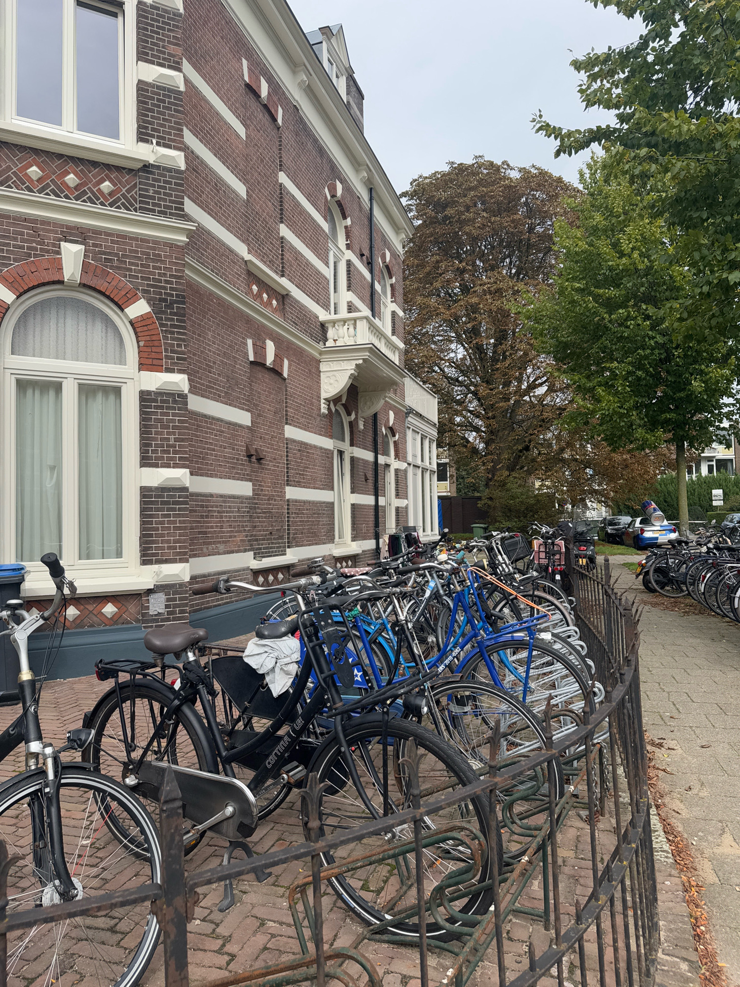 This is a medium-angle, eye-level, exterior shot of a row of parked bicycles in front of a red brick building in Nijmegen, Netherlands. The building features white trim around the windows and decorative brickwork. The sky is overcast. The bicycles are secured by a black metal rack. In the background, there are trees, a sidewalk, and a street with parked cars, including one with a police officer visible through the window. The scene appears to be an urban environment.