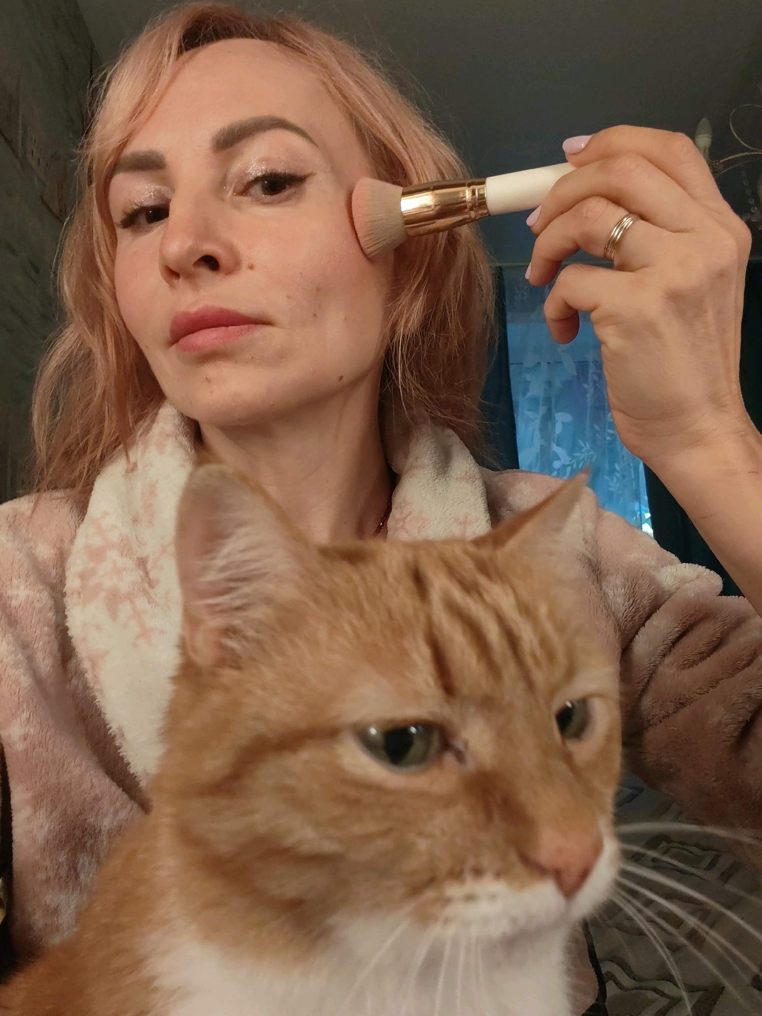 In this close-up, eye-level selfie, a woman with light pink hair is applying makeup with a fluffy brush to her cheek. She is wearing a soft, pink bathrobe with a white floral pattern. Her expression is neutral, and she has a delicate eye makeup with glitter and eyeliner. A ginger cat with green eyes is positioned in the foreground, its face filling the lower half of the frame, looking slightly off-camera. The background is dimly lit and appears to be an indoor setting with a curtain visible. The image quality is slightly grainy, suggesting it was taken with a mobile phone camera in low light conditions. The scene depicts a moment of personal grooming, with the cat as an apparent companion.