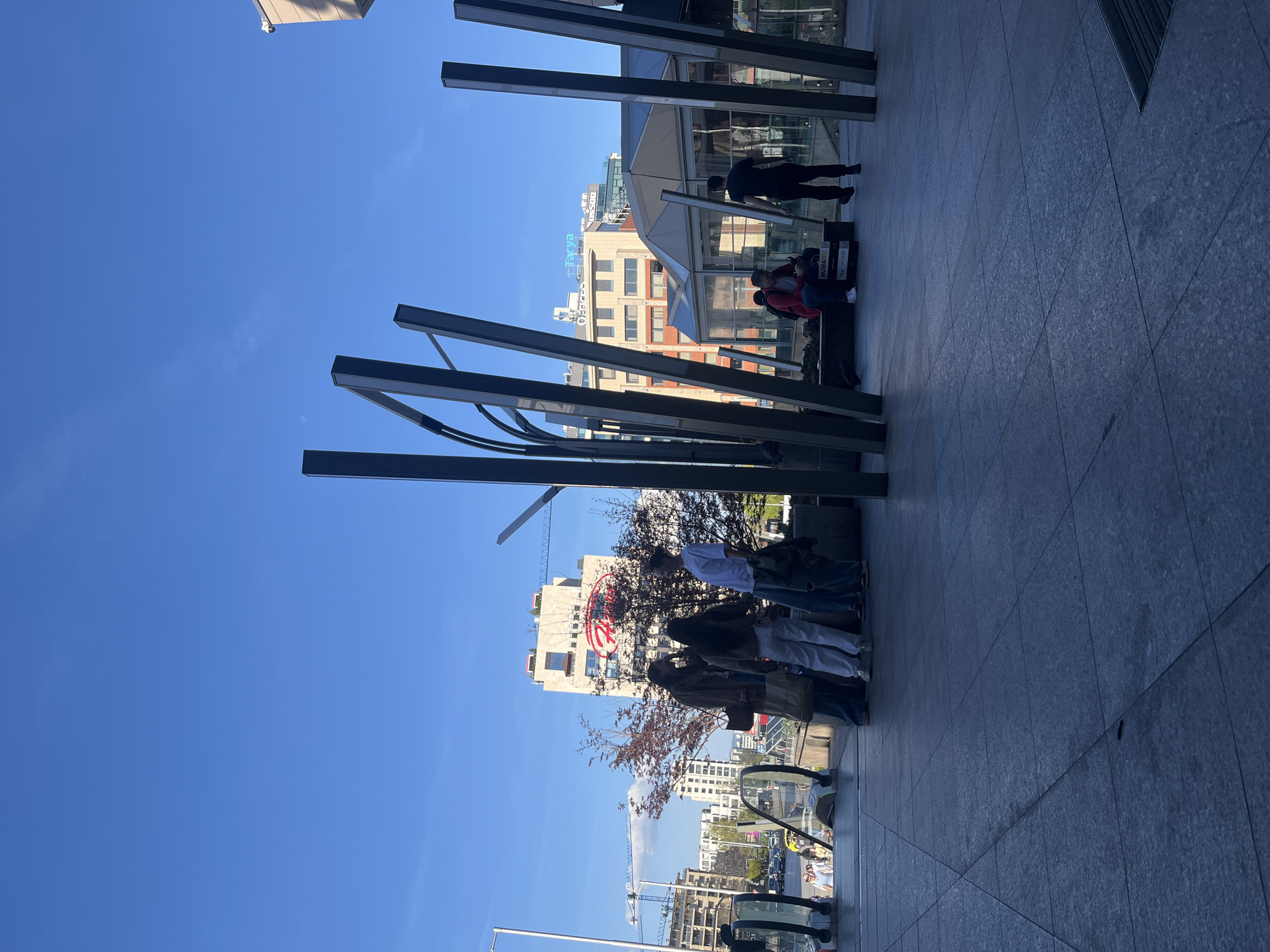 The image depicts an outdoor scene in Sofia, Bulgaria, based on visible building names (Tachya and Humana). The location appears to be a commercial area, with buildings visible in the background. Several people are present, some standing near what looks like a metal sculpture, others sitting. There is an escalator and what might be a bus station in the background. The sky is blue, and there is a construction crane visible.
