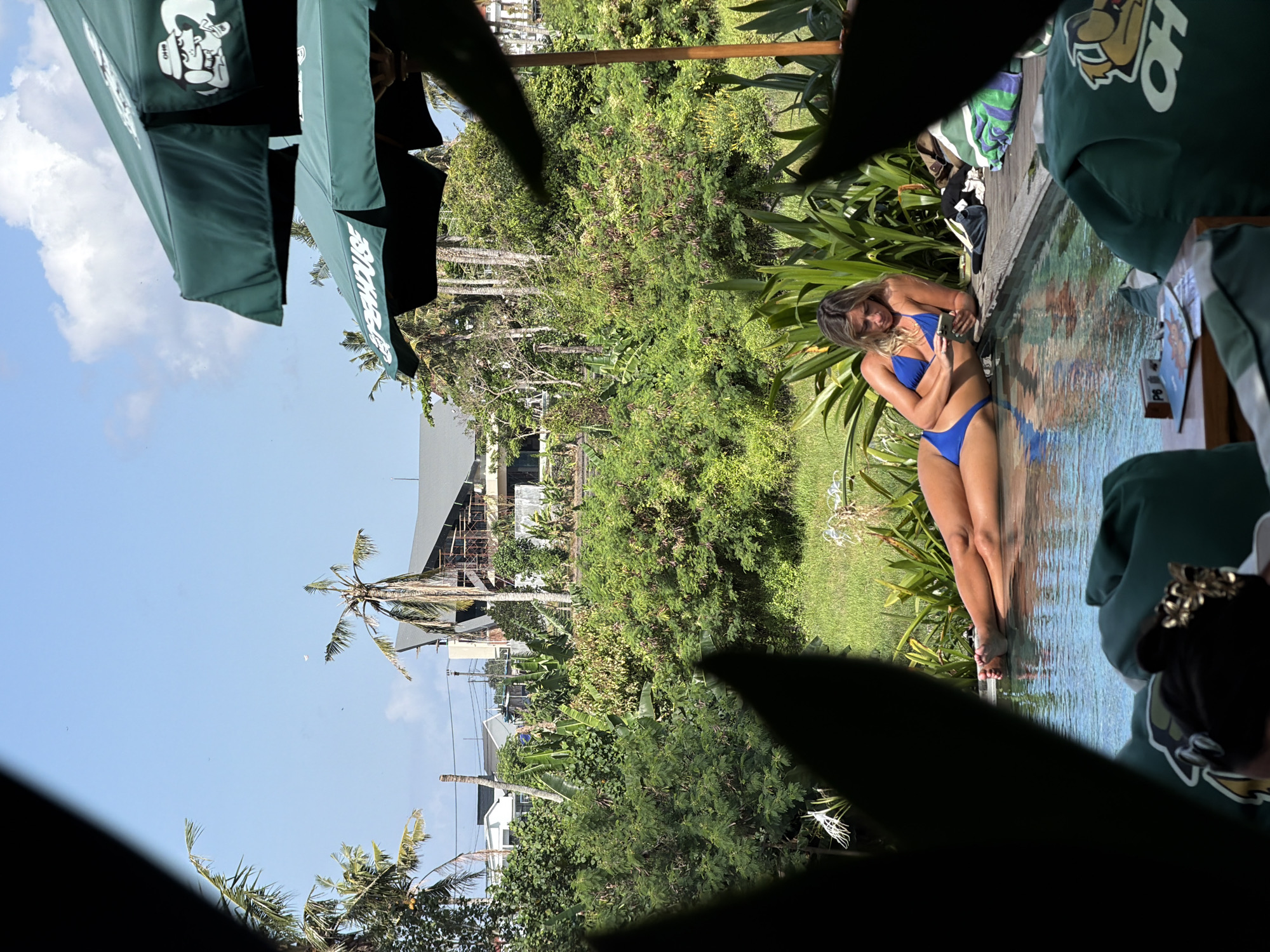 A woman in a blue bikini is lying on her side by a swimming pool, looking at her phone. She is in a lush, tropical environment with palm trees and dense green foliage. In the background, a modern building under construction is visible. Two large green umbrellas with white logos are partially obscuring the view. The scene appears to be an outdoor resort or private residence in Mengwi, Indonesia.