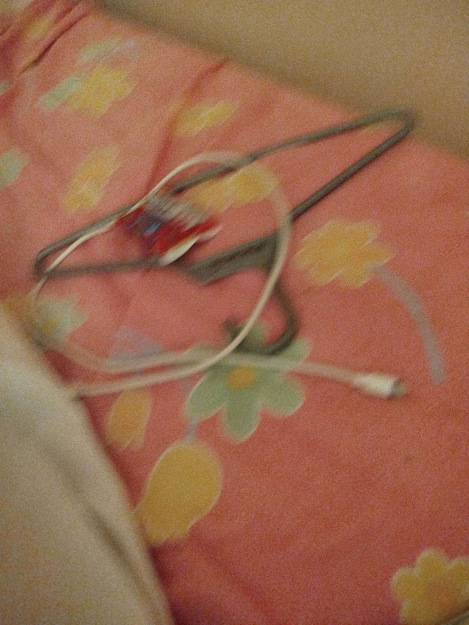 A close-up shot shows a pair of earbuds and a thin white charging cable lying on a pink fabric with a floral pattern. The earbuds are white, and the charging cable is also white, with a metallic connector. The fabric is predominantly pink with scattered yellow and light green floral shapes. The image is slightly blurry, giving it a soft focus. There is no indication of the location in Lassan, Pakistan, or any people or actions depicted.