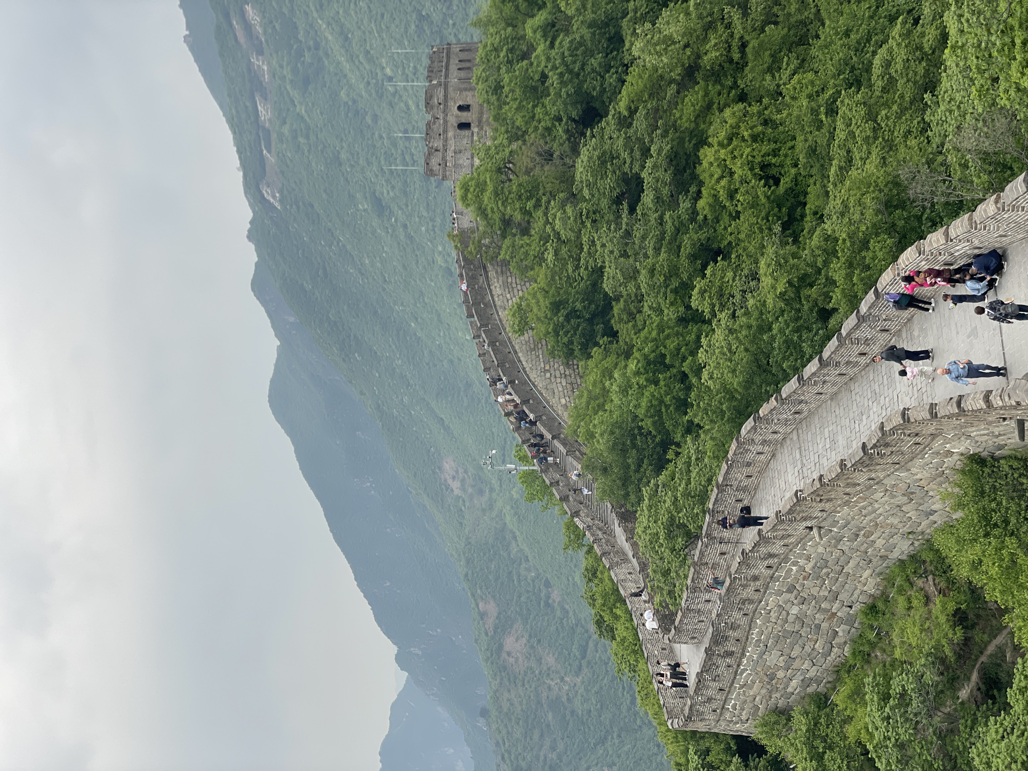 The image shows a section of the Great Wall of China in Huairou District. The stone wall winds along a mountain ridge, with lush green trees on one side and a steep mountain slope on the other. People are walking along the wall. A watchtower is visible in the distance. The sky is overcast.