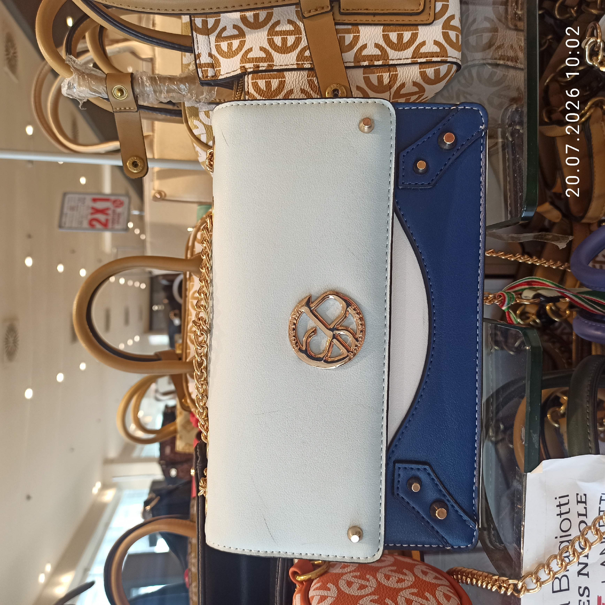 A light blue and white clutch purse with a blue bottom section is displayed on a shelf. It has a gold chain strap and a circular gold logo on the front. The purse is positioned in front of other handbags, one of which has an orange and white monogram pattern. A sign with "2X1" is visible in the background. The scene is illuminated by overhead lighting. The timestamp "20.07.2026 10:02" is present in the lower right corner, suggesting a store display. It is not possible to determine the city of Bari, Italy from the image.