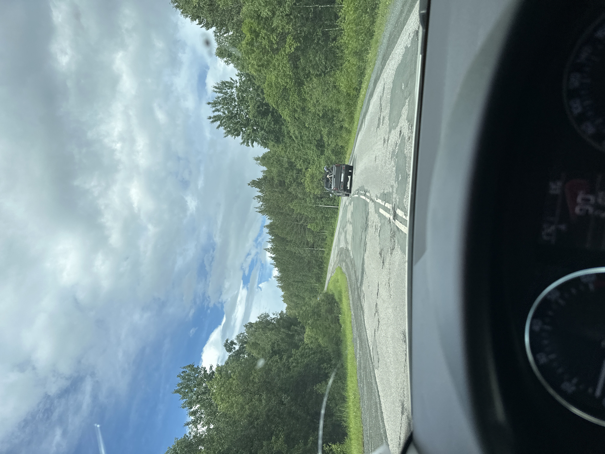 The image is a point-of-view shot from inside a car, looking out at a two-lane asphalt road. The road is in a state of disrepair with numerous potholes. A dark-colored pickup truck is visible ahead, driving in the same direction and carrying what appears to be bicycles in its bed. The road is flanked by dense green trees on both sides, forming a forest. The sky above is partly cloudy with a mix of blue sky and white clouds. The dashboard of the car is partially visible in the foreground, including a speedometer indicating a speed of 90. The scene appears to be a rural or semi-rural setting.