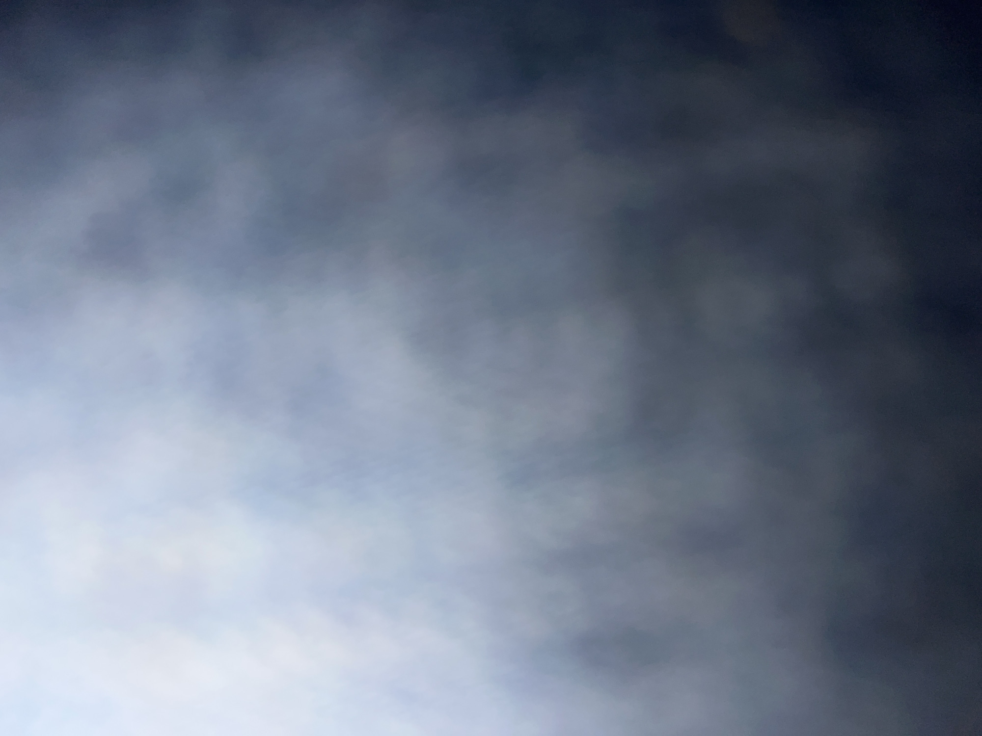 The image presents an abstract and heavily blurred composition, transitioning in color from light, hazy white and pale blue in the upper left to progressively darker shades of slate grey and deep blue towards the bottom right. The texture is soft and diffuse, suggesting a cloudy sky, dense fog, or an extremely out-of-focus background. There are no discernible subjects, objects, people, or specific actions within the frame. The lighting appears to emanate from the upper left quadrant, creating the brightest area, with the rest of the scene fading into obscurity. While the context of Casablanca, Morocco, is provided, no specific geographical or urban features of the city are identifiable in this abstract depiction.