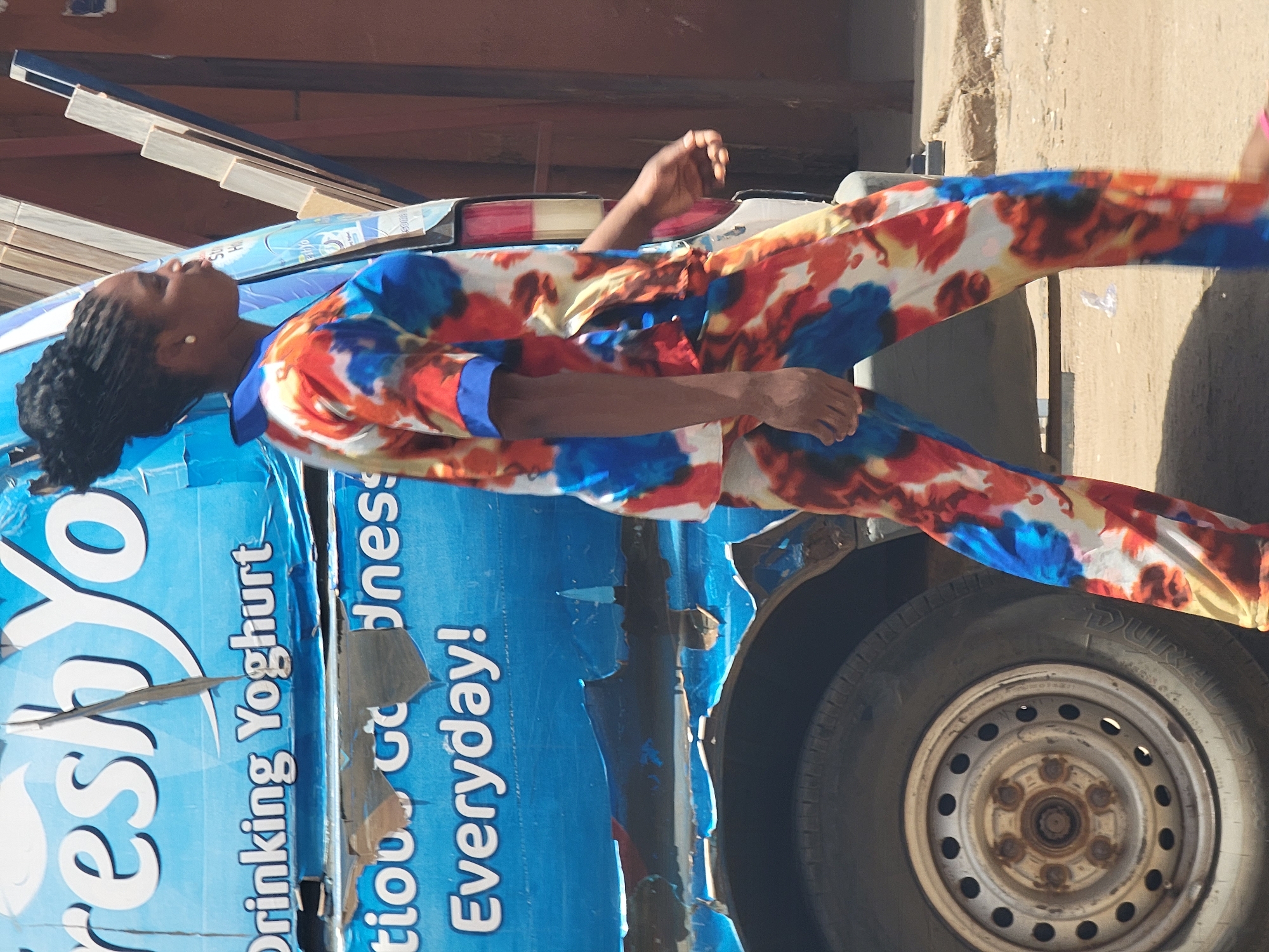 An outdoor portrait in Maiburiji, Nigeria, captures a woman standing next to a blue commercial vehicle, bathed in bright sunlight. The image is oriented with the woman's head towards the top-left and her feet towards the bottom-right.
The woman, with dark braided hair pulled back, is seen in profile, looking upwards and slightly to her right. She wears a striking two-piece outfit (a long-sleeved shirt and trousers) patterned with vibrant blue, orange, red, and white abstract or tie-dye designs. A white pearl-like earring is visible on her left ear. Her arms are relaxed, with her left hand resting near her waist and her right hand slightly lifted.
She stands beside a dilapidated blue vehicle, likely a delivery van, which is heavily branded with white text. Visible portions of the text read "Freshyo," "Drinking Yoghurt," "tion Goodness," and "Everyday!". The paint on the vehicle is visibly peeling and worn. On the vehicle's roof, what appear to be stacked, light-colored rectangular objects (possibly wooden planks or packaging materials) are visible. The rear wheel of the vehicle, with a silver-colored rim, is prominent in the bottom-right of the frame. The background includes a light-colored wall or building structure and faint shadows, indicating a clear, sunny day.