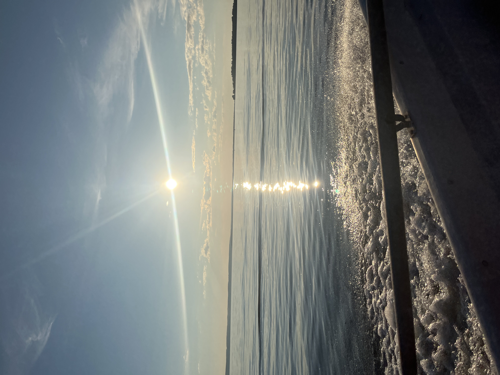 This image depicts a serene view of the wide, calm waters of Lake Ladoga near the village of Siukterka, Russia, bathed in the warm glow of the setting sun. The sun, a bright orb in the sky, casts a shimmering reflection across the water's surface, creating a dazzling path of light. Gentle ripples disturb the water, suggesting a slow-moving vessel, possibly a boat or a ferry, as evidenced by the foamy wake visible in the foreground. A metallic railing, likely part of the boat's structure, runs horizontally across the bottom of the frame, adding a sense of perspective and hinting at the viewer's vantage point. In the distance, a low, tree-lined horizon marks the distant shore. The sky is a pale blue, transitioning to a warmer hue near the horizon, with a few wispy clouds adding texture to the scene. The overall impression is one of tranquility and natural beauty, characteristic of a peaceful day on the lake.
