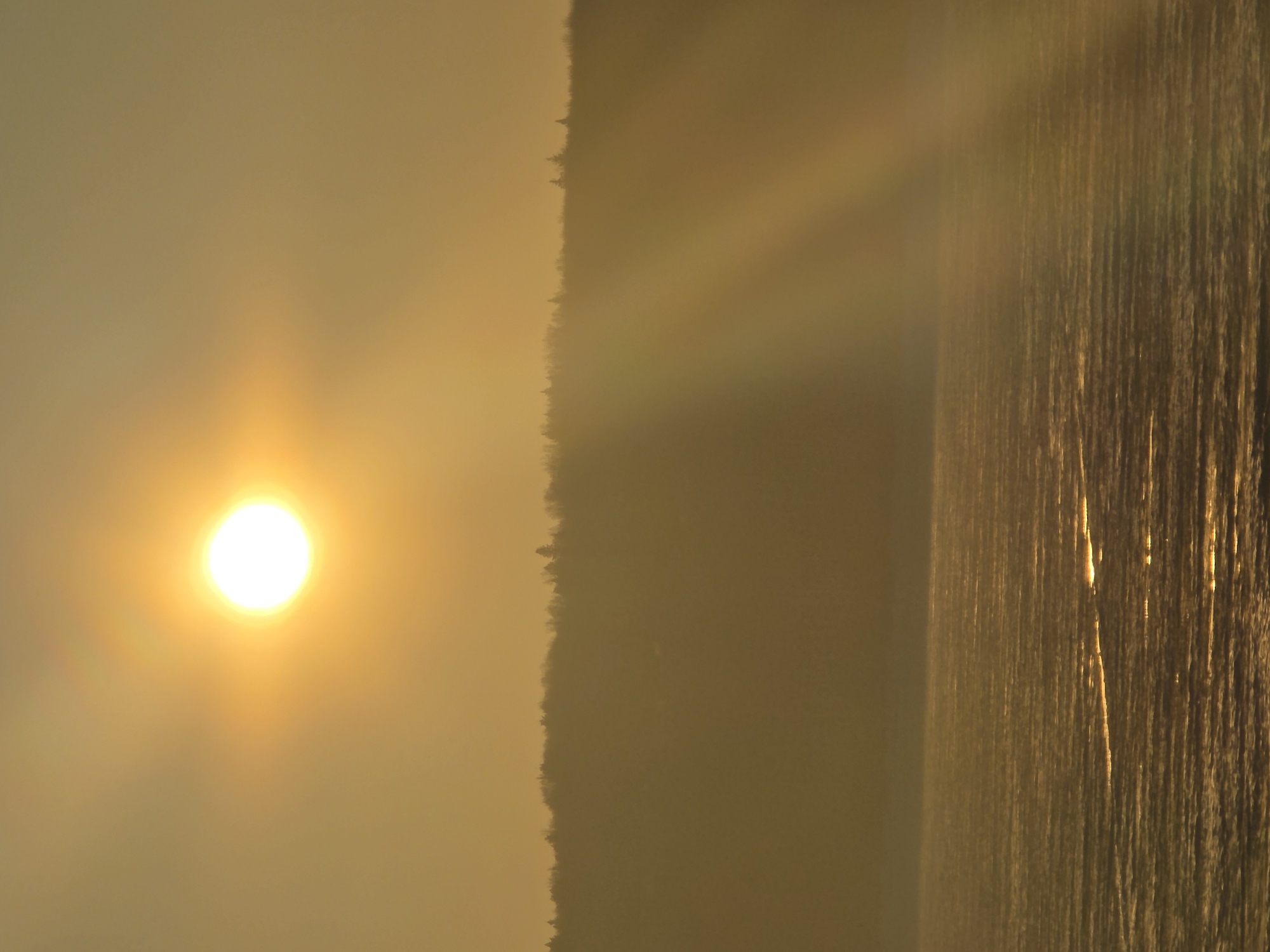 The image depicts a bright sun in the sky over the city of Chaikovsky, Russia, with a vertical structure or waterfall illuminated by sunlight on the right side of the frame. A dark silhouette of trees or a building is visible between the sun and the illuminated structure. The environment appears hazy or foggy.