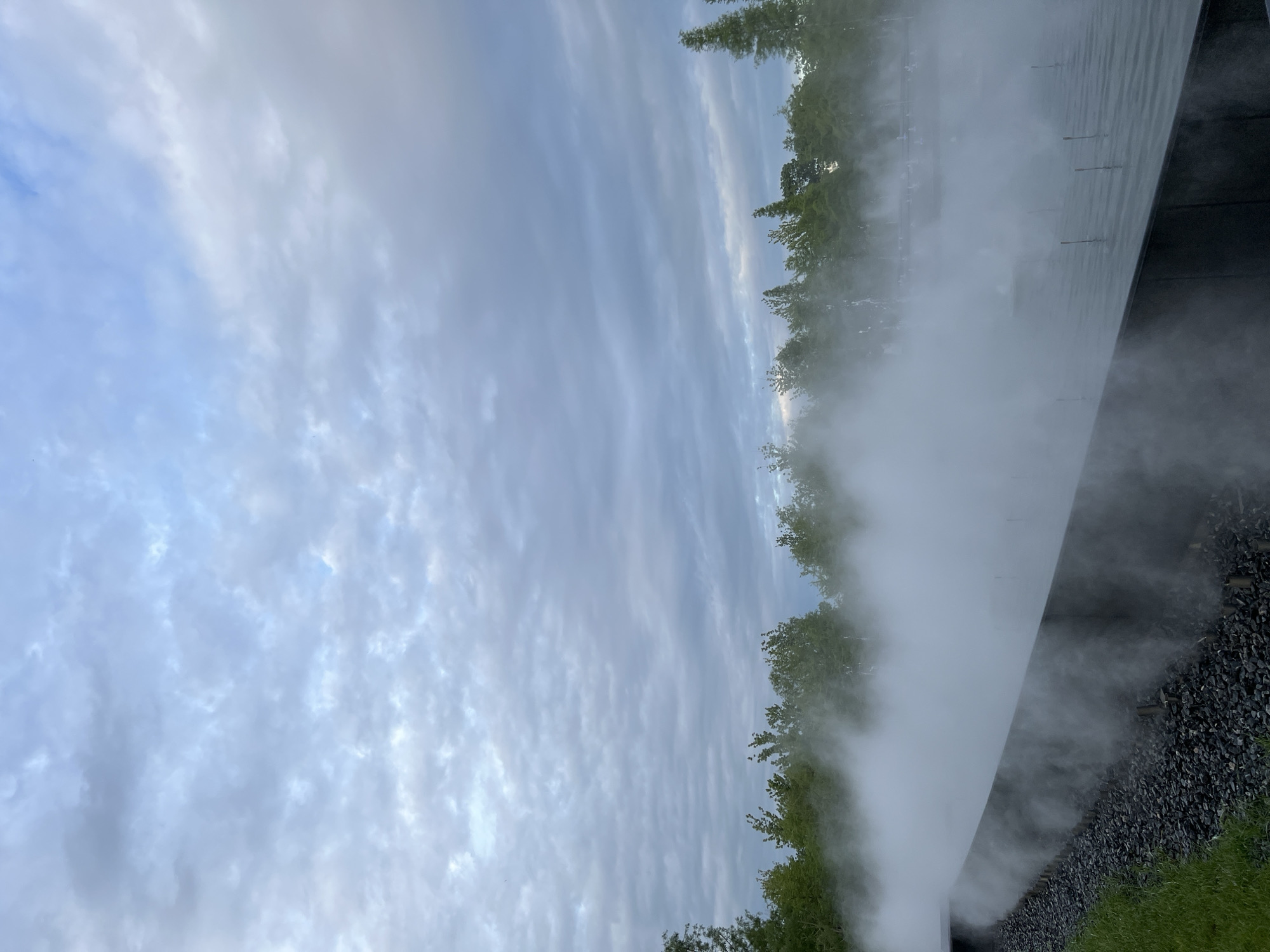 The image shows a sky filled with streaky clouds, juxtaposed with a structure emitting a plume of white mist along its base. The lower part of the structure sits atop dark gravel and a strip of green grass. Trees are partially obscured by the mist. The location is not discernible from the image alone.