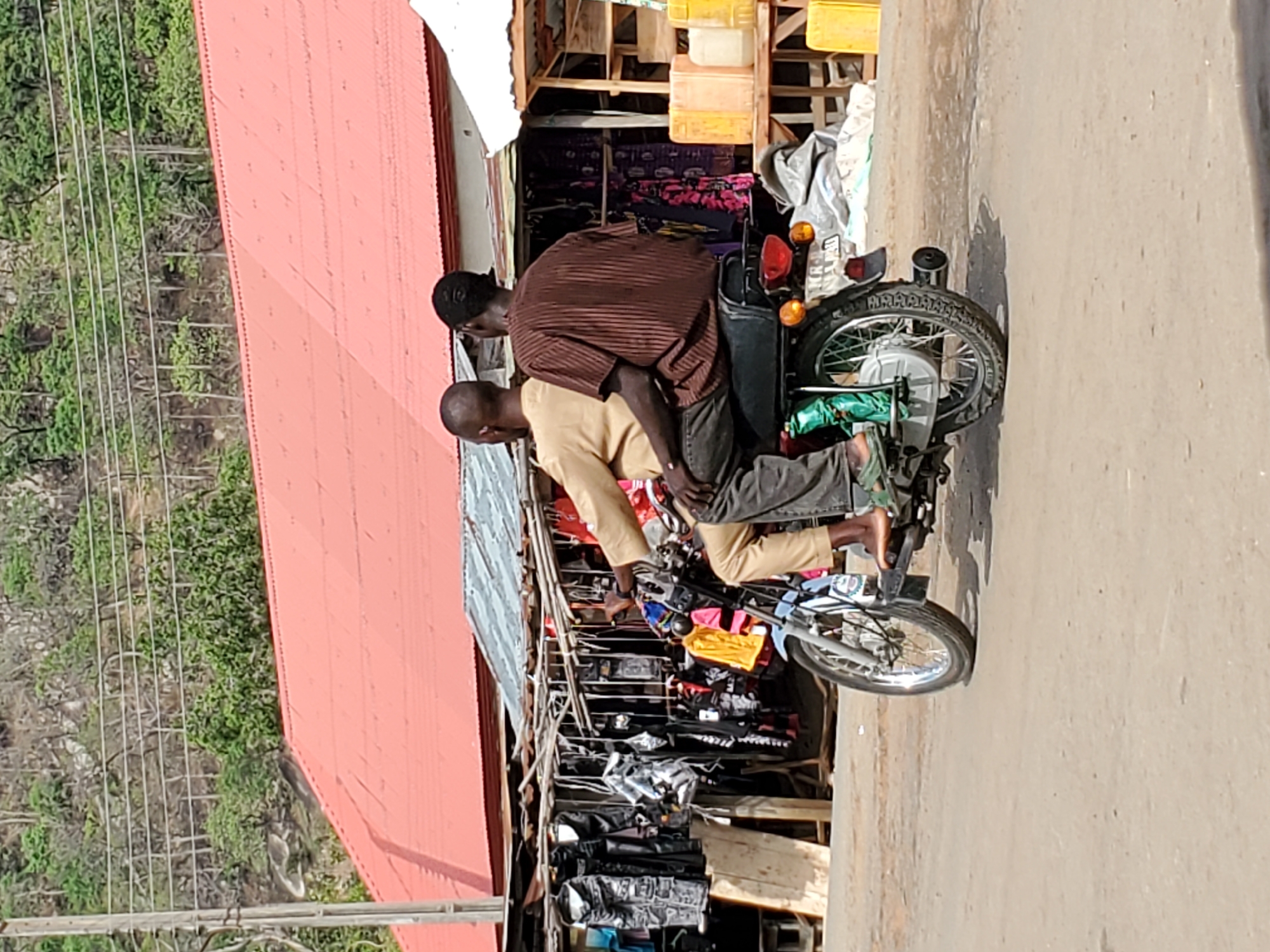 Two men are on a motorbike parked in front of a shop in Mararaba, Nigeria. The shop sells clothes. There is a red roof building in the background. A mountain can be seen in the distance.