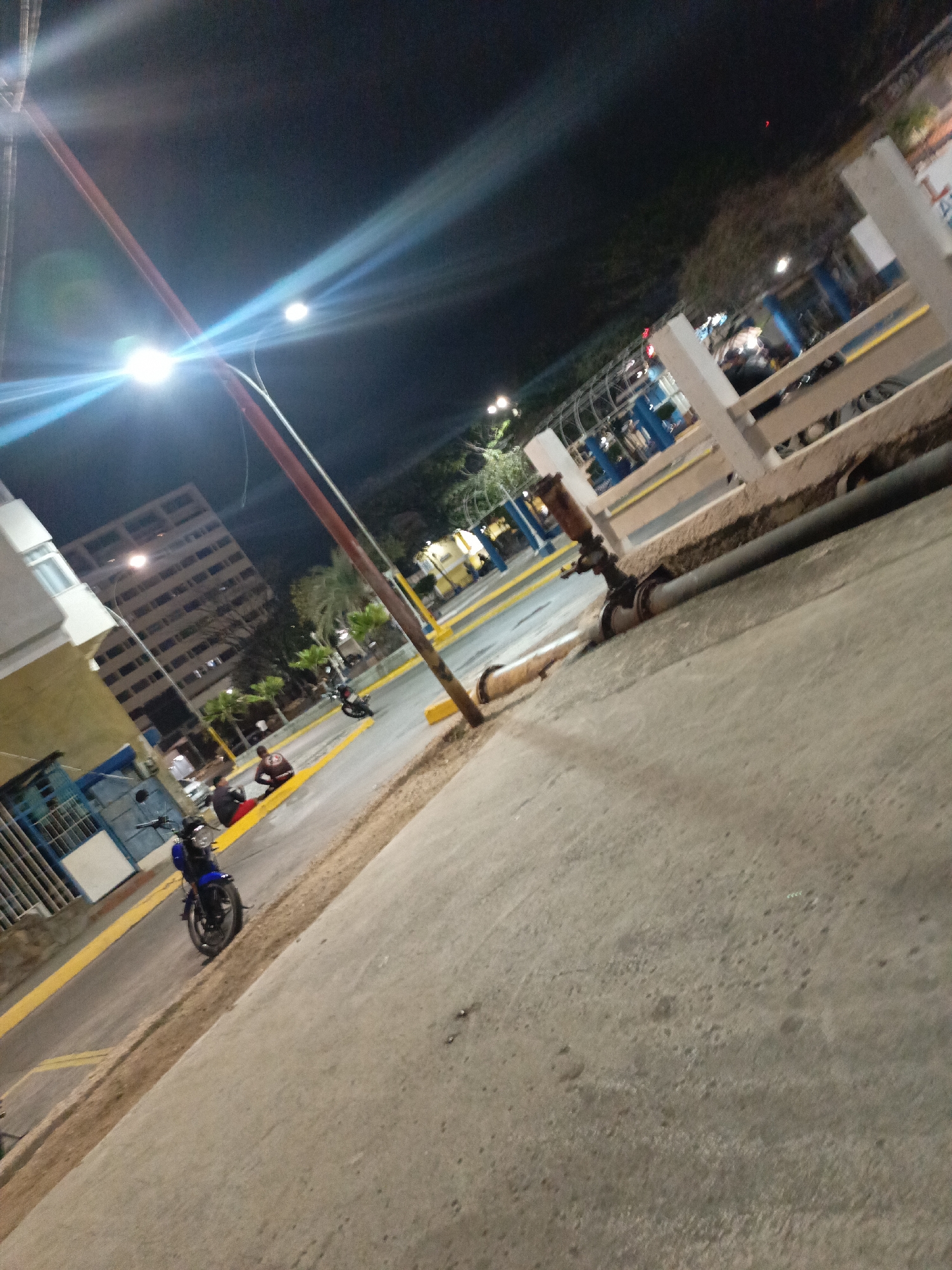 The image shows a street scene in Juan Griego, Venezuela at night. A street light casts a bright glare, and the environment appears to be urban. A yellow curb with painted road stripes divides the street from the sidewalk. A blue motorcycle is parked on the roadside. Two people are seated near the curb. A large building with multiple windows is visible. A rusted pipe runs along the sidewalk near a painted fence. Trees and shrubs are in the background.