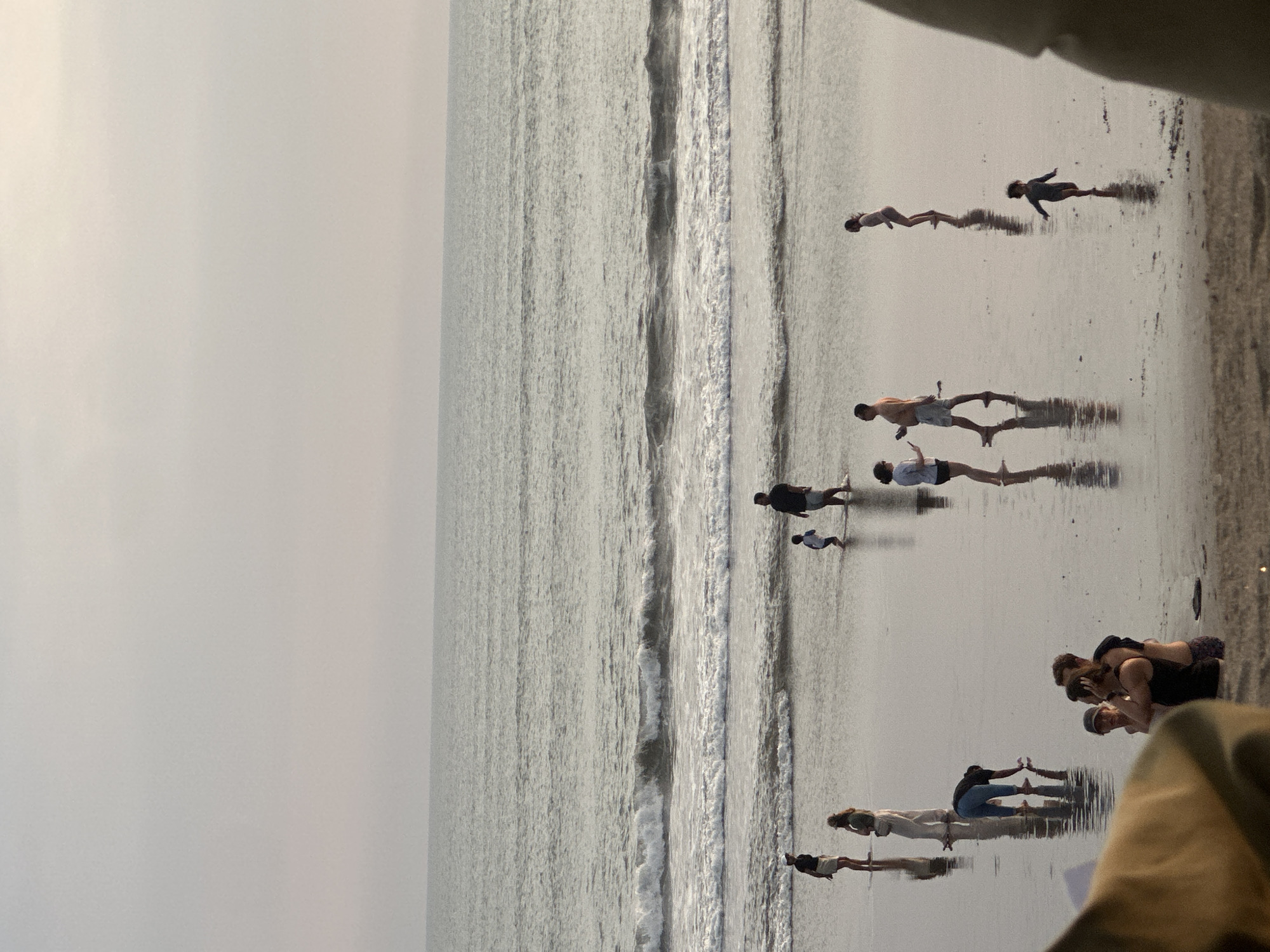 In North Kuta, Indonesia, a beach scene unfolds with several individuals scattered across the wet sand and shallow water. The sky is overcast and hazy, with a muted light illuminating the ocean. Gentle waves break near the shore, creating ripples that reflect the figures on the beach.
To the left, a person in a white hat and dark shirt stands facing the water, while another person in a light-colored outfit walks away. Further down the beach, a person in blue pants bends over, touching the wet sand. Near the center, a couple embraces, with a child partially visible between them. To their right, a man in a black shirt stands with his back to the viewer, while a small child runs towards the water. A woman in a light blue shirt and dark shorts walks to the right, and a woman in a pink swimsuit stands further out in the water. In the foreground, a large, out-of-focus object, possibly a piece of furniture or fabric, partially obstructs the view.