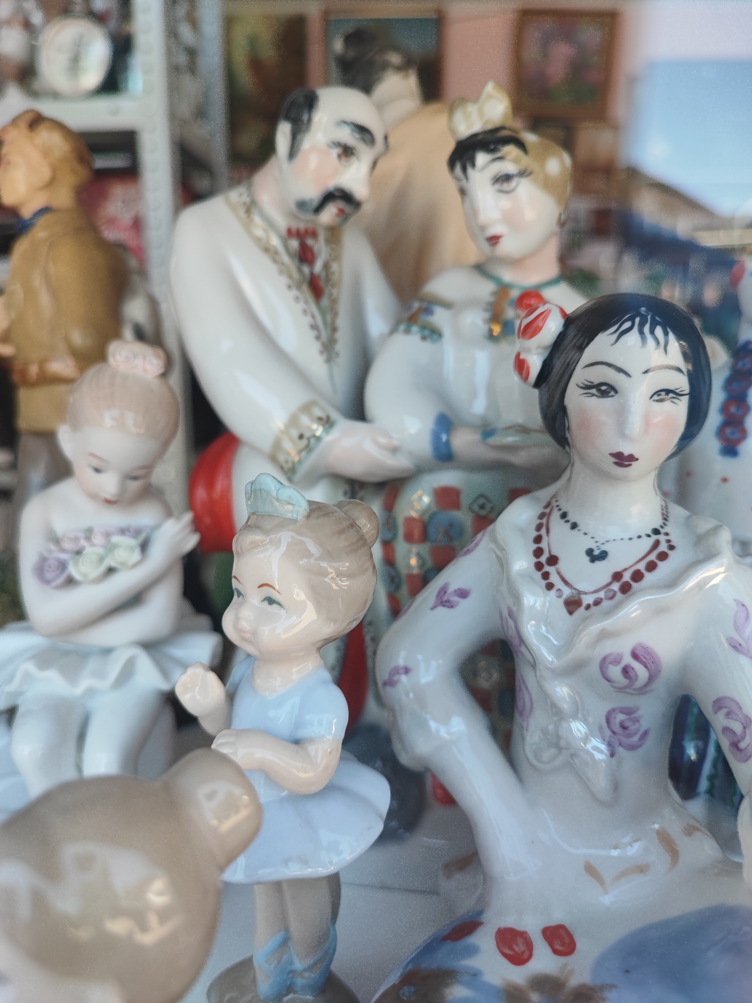 This is a close-up, eye-level shot of a collection of porcelain figurines on display, possibly in a shop or home in Pyatigorsk, Russia. The figurines depict a variety of characters, including what appears to be a couple in traditional attire, several young girls dressed as ballerinas, and a neutral-colored, possibly animal figurine in the foreground. The central couple is dressed in white, with the man sporting a mustache and the woman wearing a headdress and ornate clothing. The ballerinas are delicate, with one holding a bouquet and another in a tutù. The figurines are arranged in a densely packed display, with some partially obscured by others. The background is out of focus, suggesting shelves or other display elements. The lighting is soft, and the overall impression is one of an artistic or collectible arrangement.