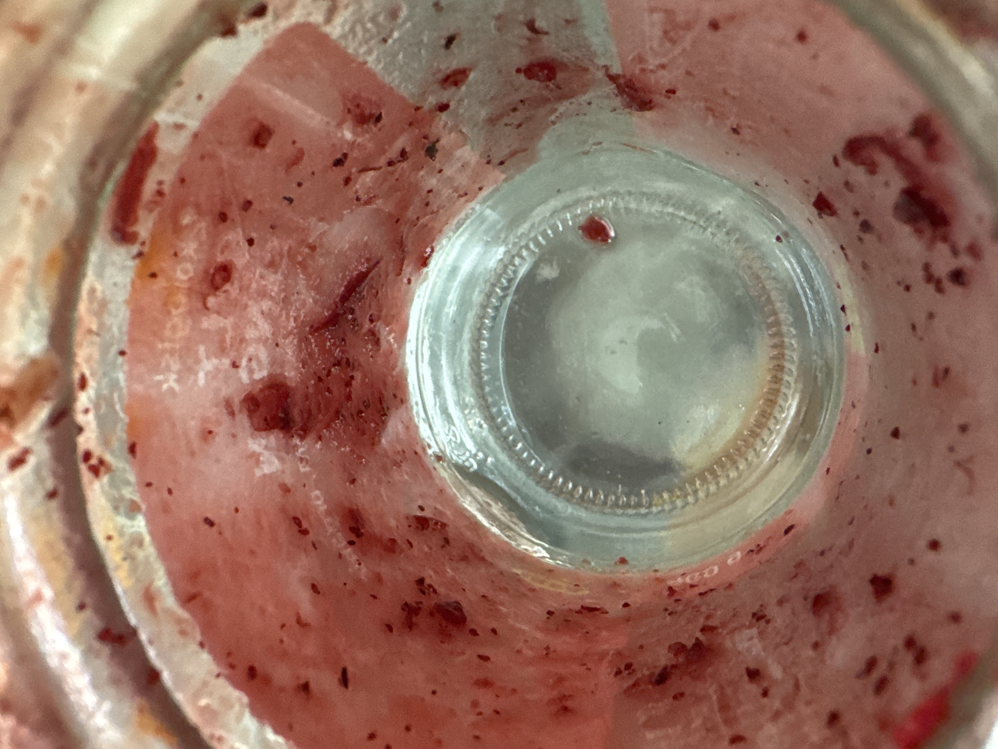 This is a close-up, overhead shot looking down into a glass jar that has residue from a red, likely fruit-based, substance. The inside of the jar is coated with pinkish-red pulp and small dark specks, consistent with fruit remnants. The bottom of the jar is visible as a clear, circular glass surface with a ribbed texture around the edge. The lighting suggests an indoor environment, possibly a kitchen or dining area. There is no information available to link this image to the city of Rawai, Thailand.