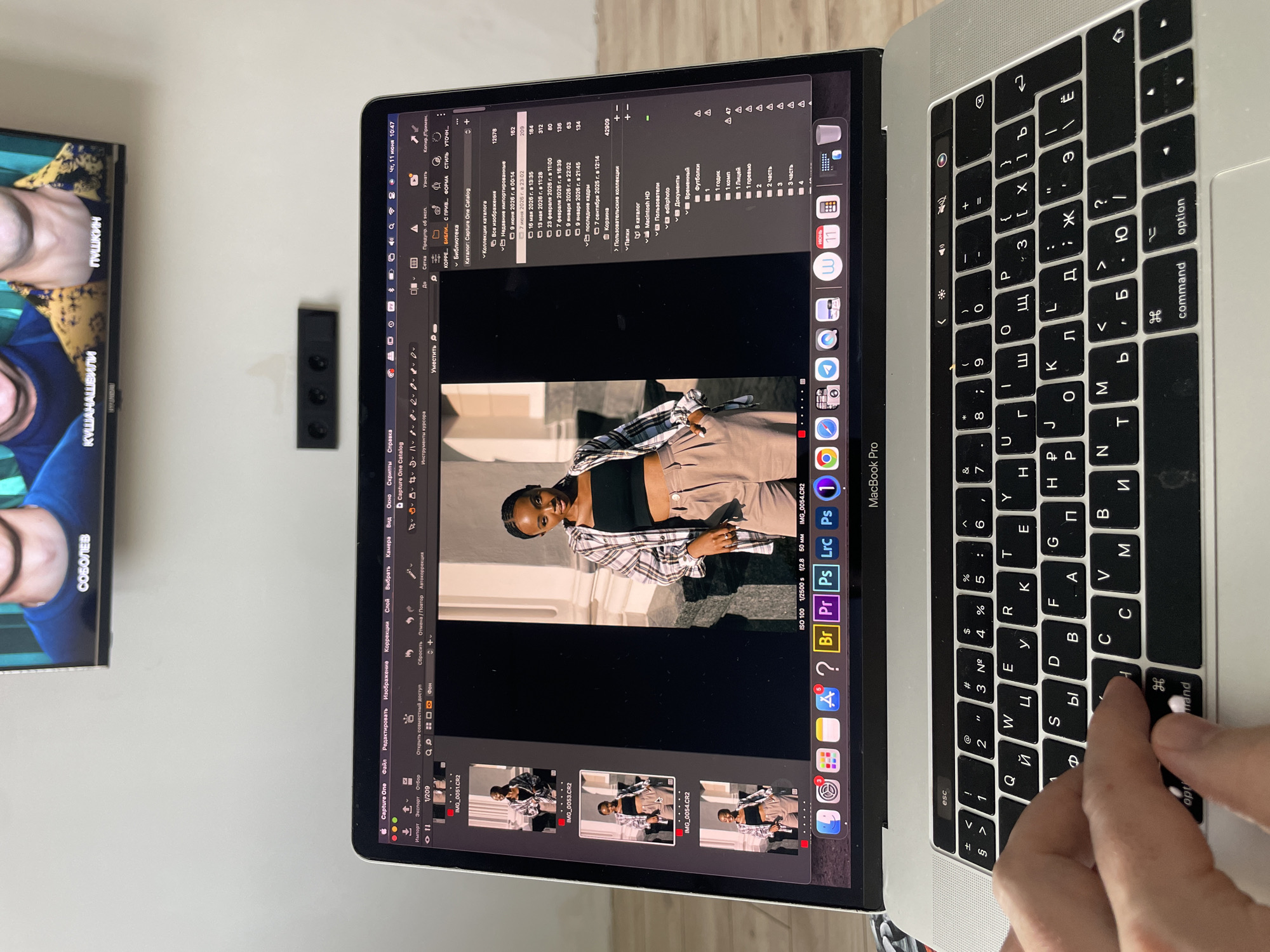 The image shows a person's hands on a MacBook Pro laptop, with the screen displaying the Capture One photo editing software. The software is showing a portrait of a young Black woman wearing a plaid shirt and tan pants, with other similar photos in a thumbnail view to the left. The laptop is open and placed on a surface, likely a table or desk, with a portion of the keyboard visible. In the background, a television screen displays a picture of three men, with Russian text below them that reads "СОБОЛЕВ" (Sobolev), "КУШАНАШВИЛИ" (Kushanashvili), and "ПУШКИН" (Pushkin). Below the TV, a black electrical outlet strip is mounted on a white wall. The lighting suggests an indoor environment. There is no indication that this scene is located in Kazan, Russia, or any specific city.
