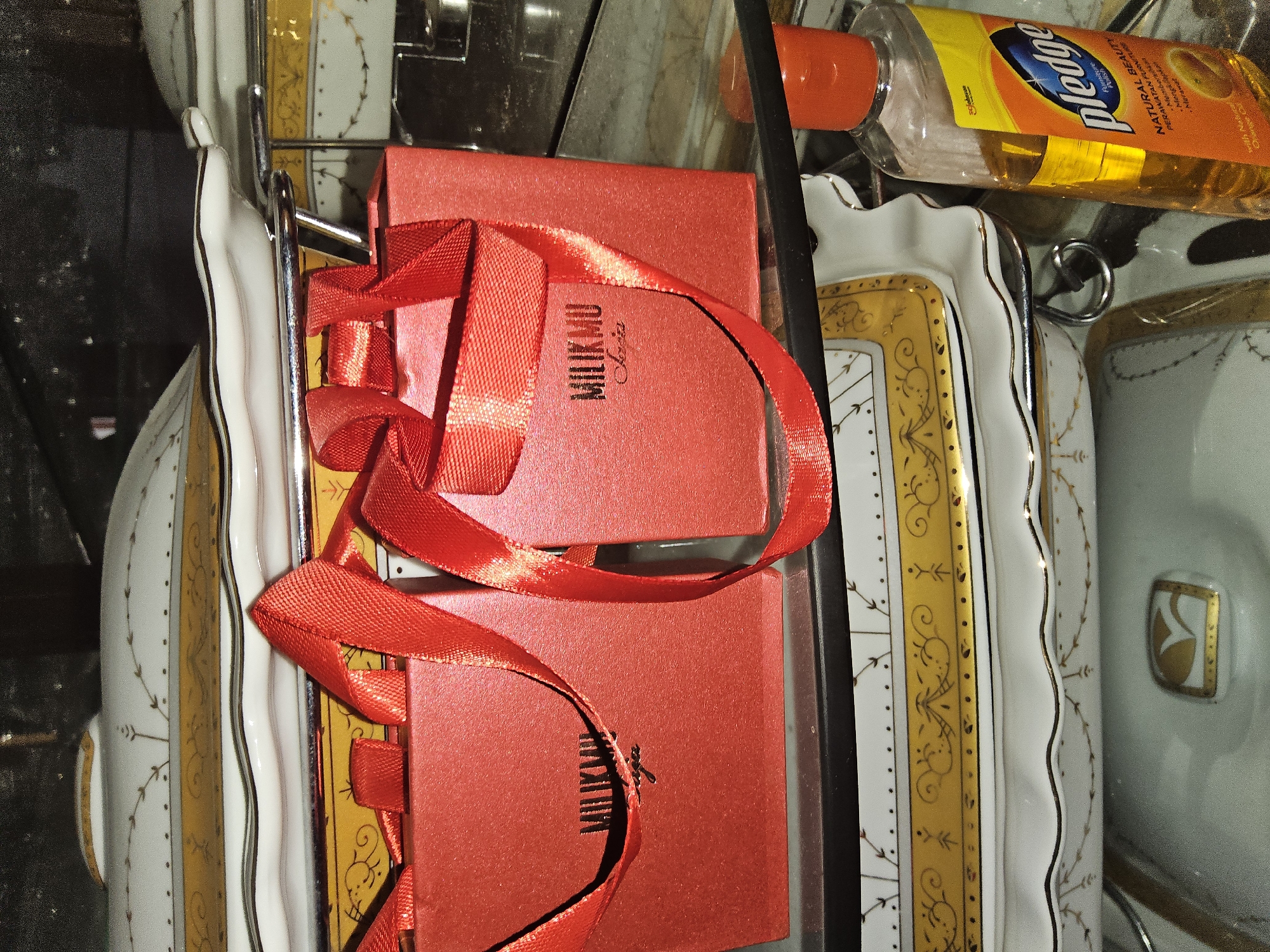 A bottle of Pledge furniture polish, with an orange cap and a yellow and orange label, stands on a glass surface next to two small, red gift boxes tied with red ribbons. The gift boxes have the word "MILIKMU" embossed on them. Behind the gift boxes and bottles are stacked ceramic serving dishes with gold trim. The background is dark and out of focus. The scene is located in a retail setting, possibly a store in Payagaboh, Indonesia, given the presence of the Pledge furniture polish which is a common household product. There are no people visible in the image.