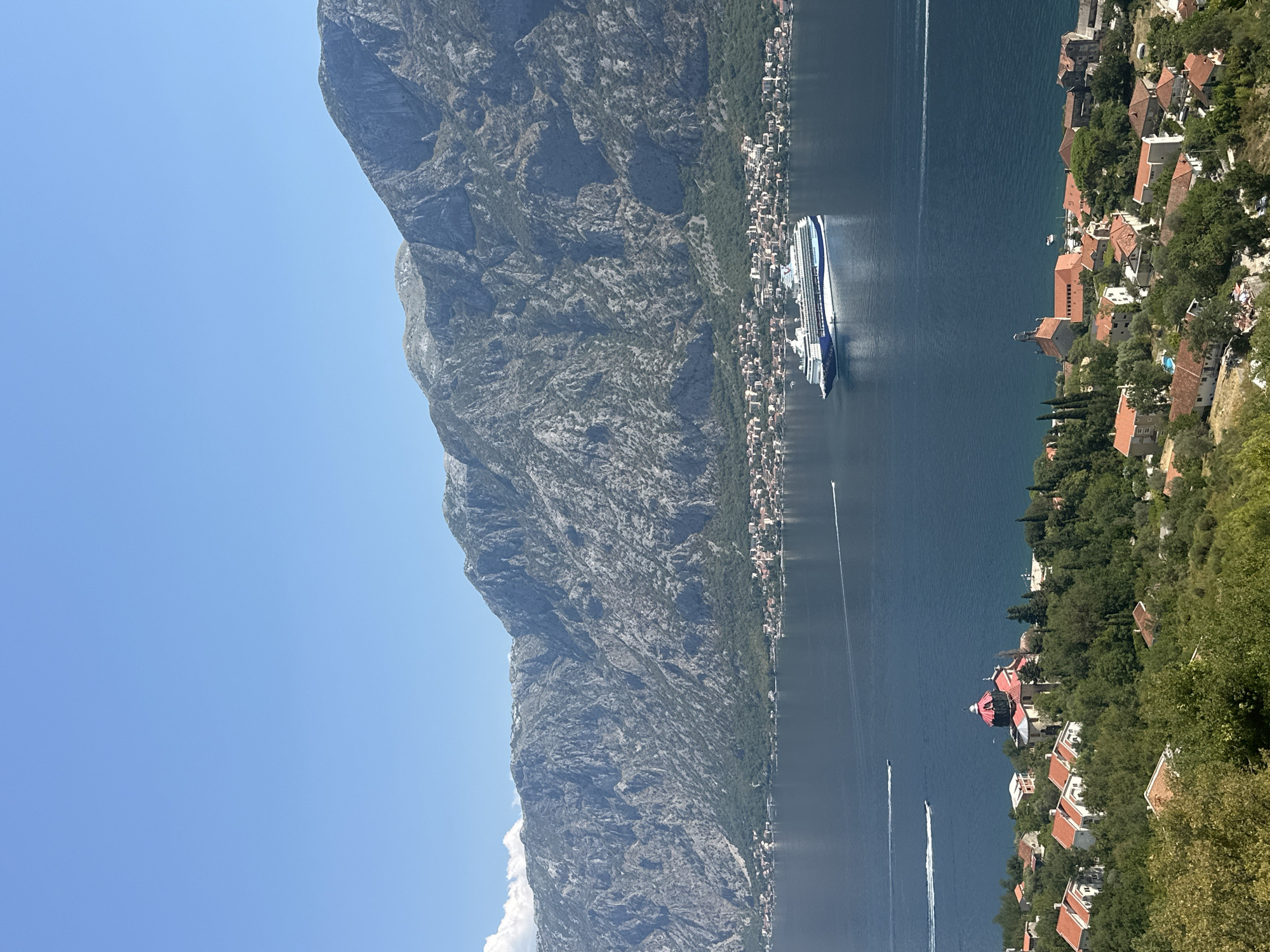 This is an eye-level, medium shot photograph taken from a high vantage point overlooking a coastal town. The scene is dominated by a large, blue body of water, likely a bay or fjord, leading to a mountainous backdrop. A large cruise ship, predominantly white with blue accents, is docked or moving slowly on the water. Several smaller boats are also visible on the water, leaving wakes.
In the foreground and middle ground, a densely populated coastal settlement is visible, characterized by red-tiled roofs and buildings interspersed with lush green trees. One prominent building, possibly a church, stands out with a red dome. The settlement extends along the coastline, with the base of the imposing, rugged mountains forming the far backdrop. The sky is a clear, bright blue with a few wispy clouds. The image is captured on a sunny day, as indicated by the strong lighting and clear visibility. The scene is consistent with the landscape of Dobrota, Montenegro, known for its bay, mountains, and historic settlements. There are no people or animals clearly visible in the frame.