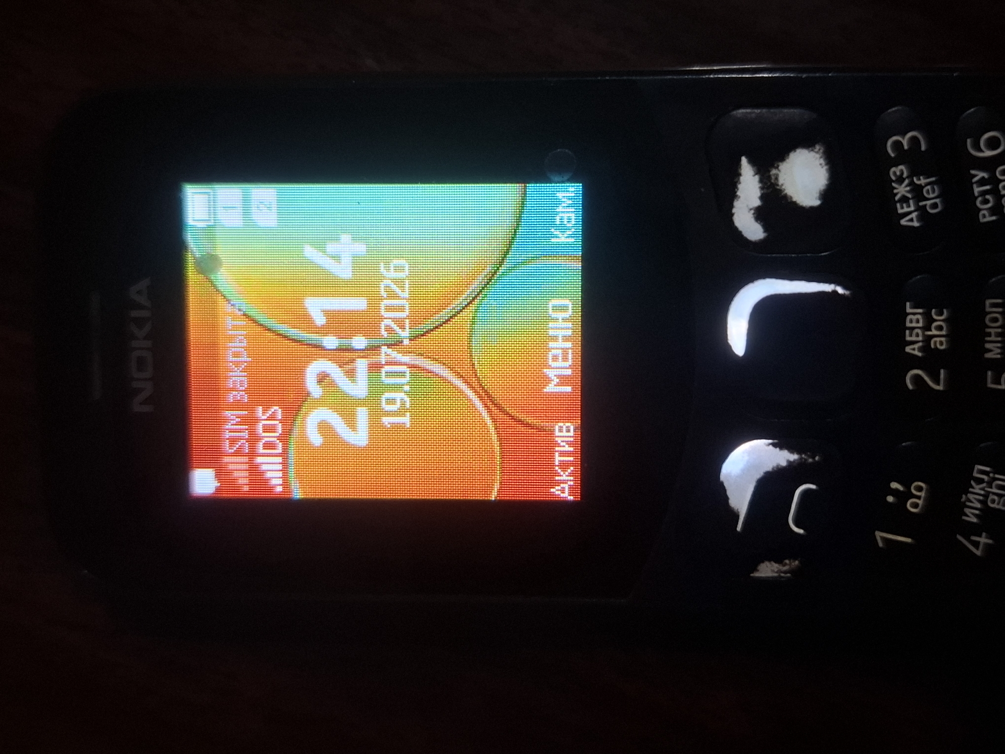 The image shows a close-up of a black Nokia mobile phone screen displaying the time 22:14 and the date 19.07.2026. The screen also shows signal strength indicators, battery level, and text labels for menu options: "АКТИВ" (Active), "Меню" (Menu), and "Кам." (Camera). The phone is dark with a brightly lit screen. Below the screen, the keypad is visible with the numbers 1 through 9 and corresponding letters. Some of the buttons have a bright, reflective surface, possibly from ambient light or a flash. The background is dark and appears to be indoors. There is no identifiable information or context in the image to suggest it is located in Almaty, Kazakhstan.