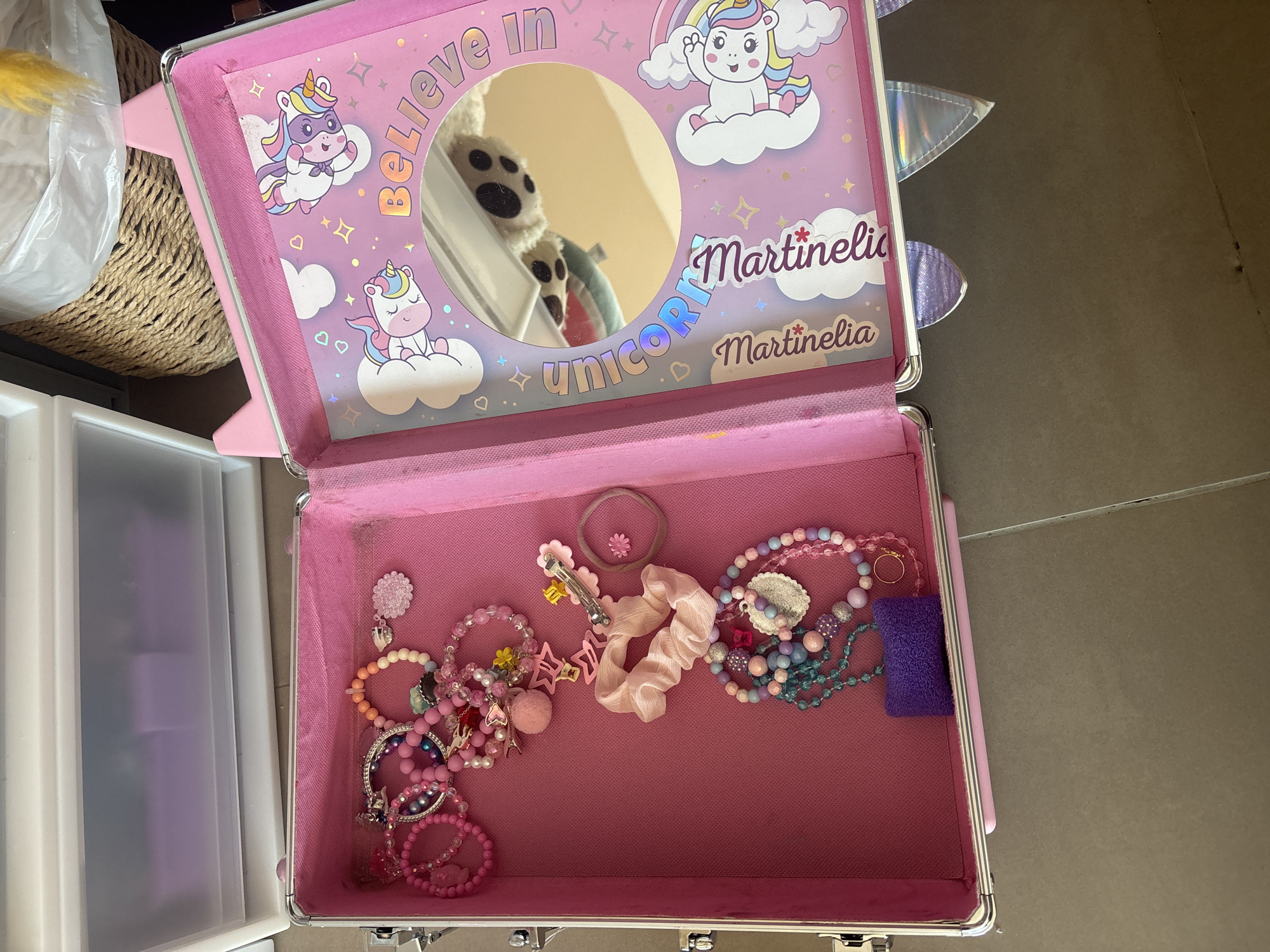 The image shows an open pink jewelry box with various accessories inside, including bracelets, necklaces, hair clips, and scrunchies. The interior of the lid is decorated with unicorn-themed graphics and a mirror. The jewelry box is placed on a gray tiled floor next to a white plastic drawer unit and a woven basket. The scene appears to be indoors, possibly in a home. There is no indication of the location being in Dubai, United Arab Emirates.