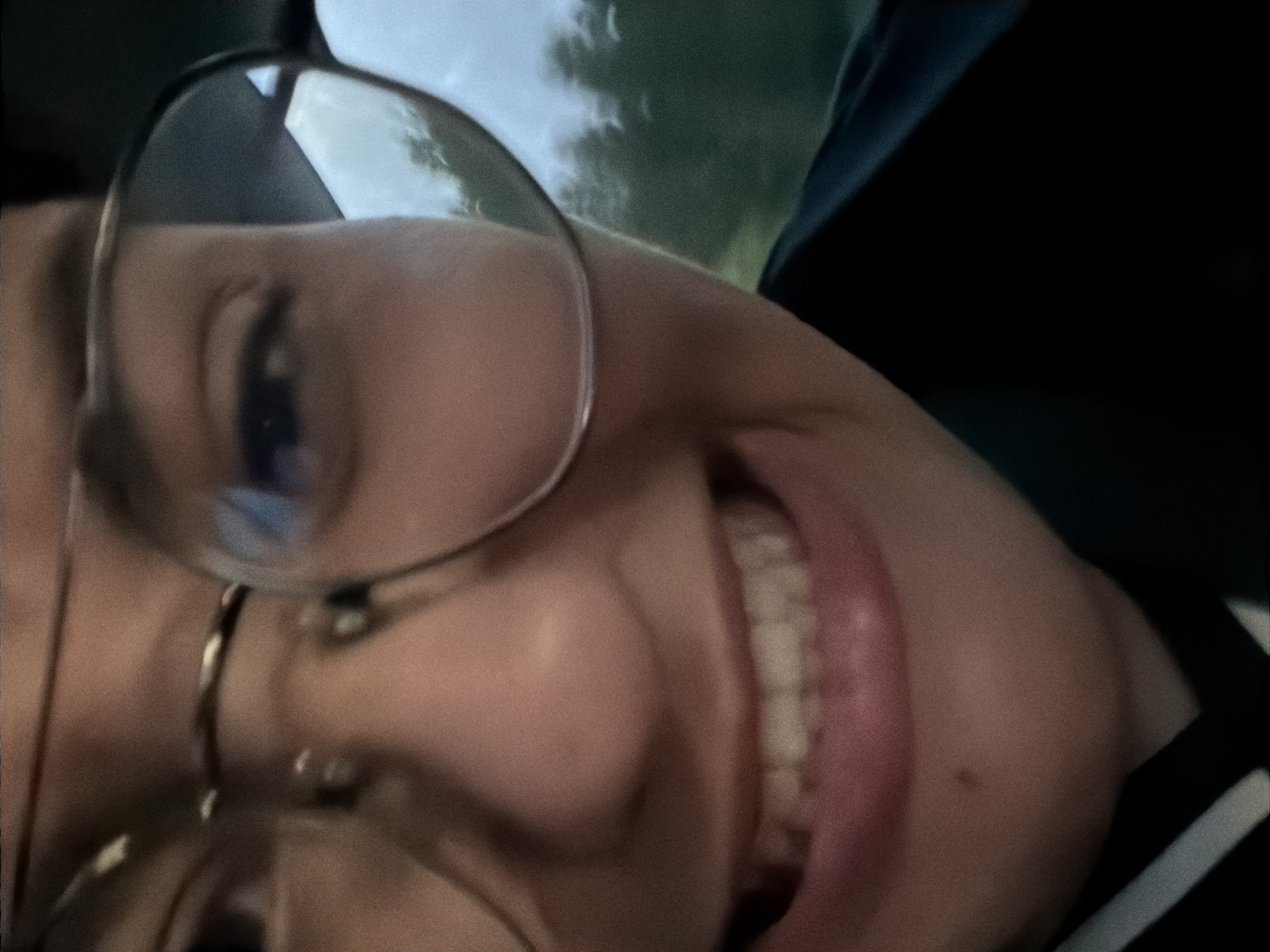A close-up, slightly blurry, selfie-style photo shows a person's face from the chest up, with a smiling expression. The person is wearing large, round glasses. The background is out of focus and appears to be a natural landscape with trees and a cloudy sky, suggesting an outdoor setting. The exact location of Rygge, Norway is not discernible from the visual elements within the image.