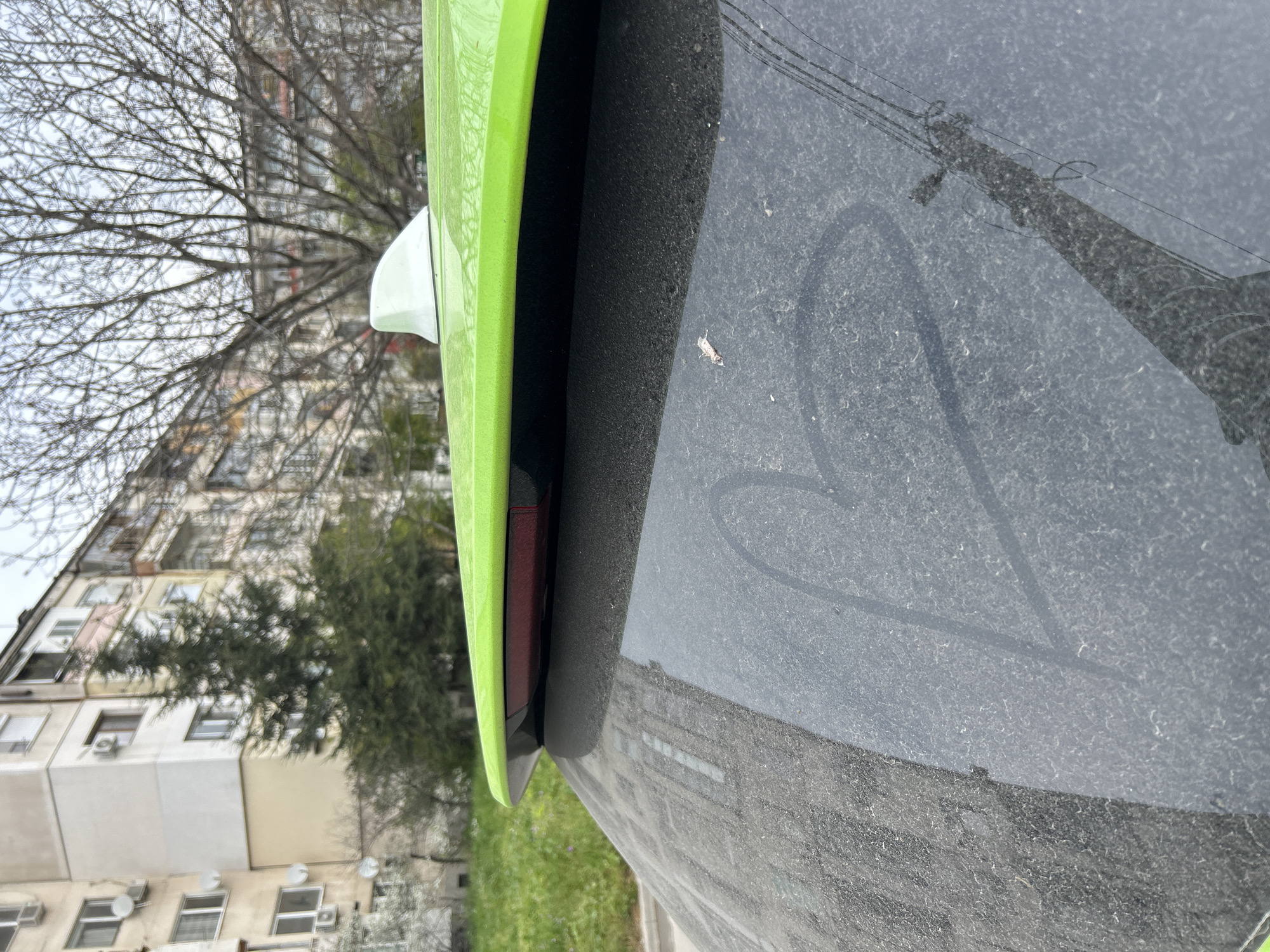 The image shows the rear of a lime green car, heavily coated in dust. A heart has been drawn in the dust on the rear surface. The car is parked on a street in Plovdiv, Bulgaria. In the background, there is a multi-story residential building with trees partially obscuring it. A utility pole is reflected in the car's dust-covered surface.