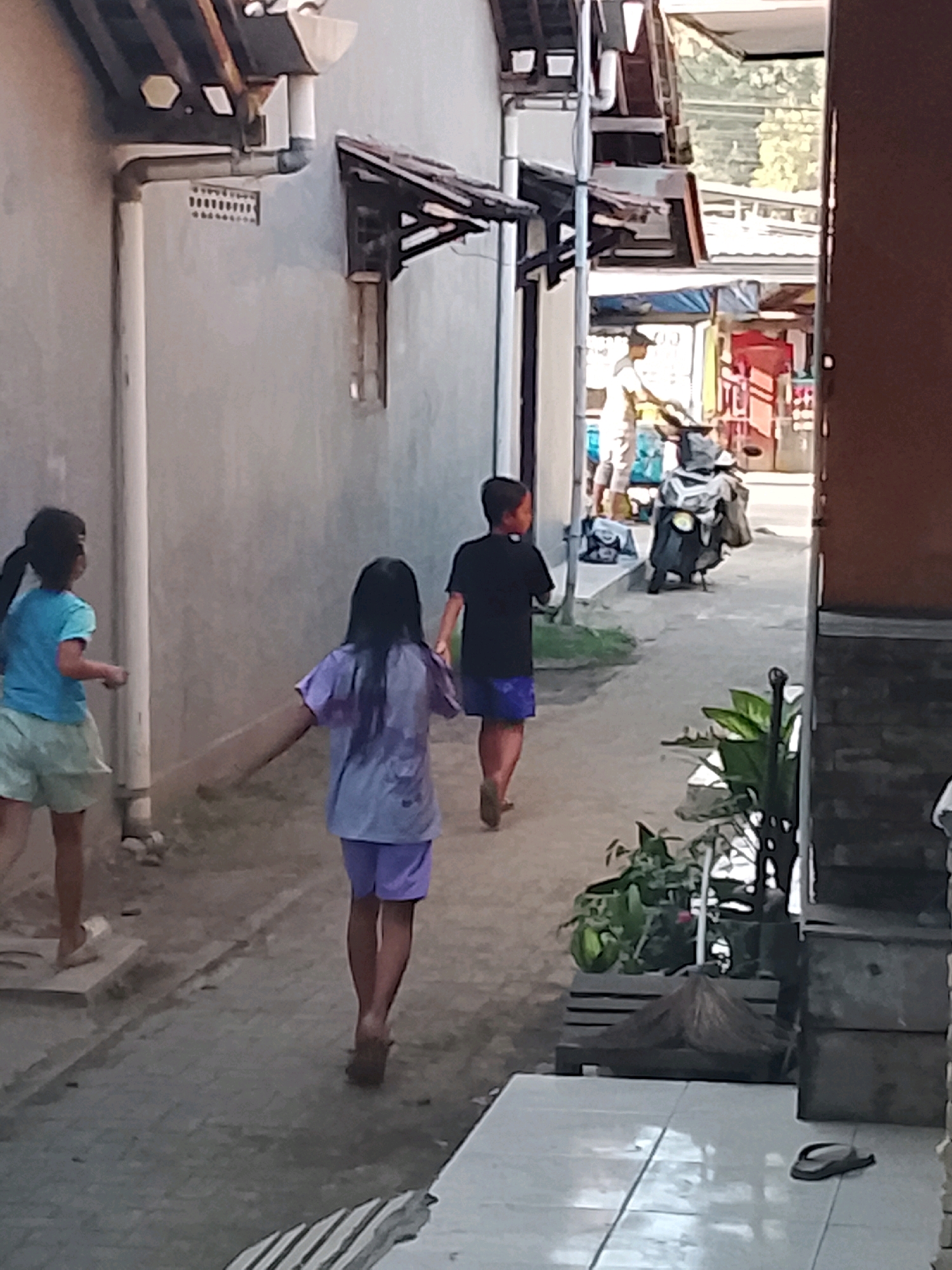 In the city of Ujungrusi, Indonesia, a group of children are seen walking down a narrow, paved alleyway. A young girl in a blue t-shirt and light green shorts is on the left, facing away from the camera. To her right and slightly behind her, another young girl with long dark hair, wearing a purple t-shirt and light purple shorts, is walking away from the camera. A young boy in a black t-shirt and blue shorts is walking further down the alley, also facing away. A parked motorcycle with a gray cover is visible in the background on the right side of the alley. The alleyway is flanked by buildings with gray walls and tiled roofs. There are some plants in pots on the right side of the alley, and a pair of flip-flops rests on the tiled ground in the foreground. The lighting suggests it is daytime with sunlight filtering into the alley.