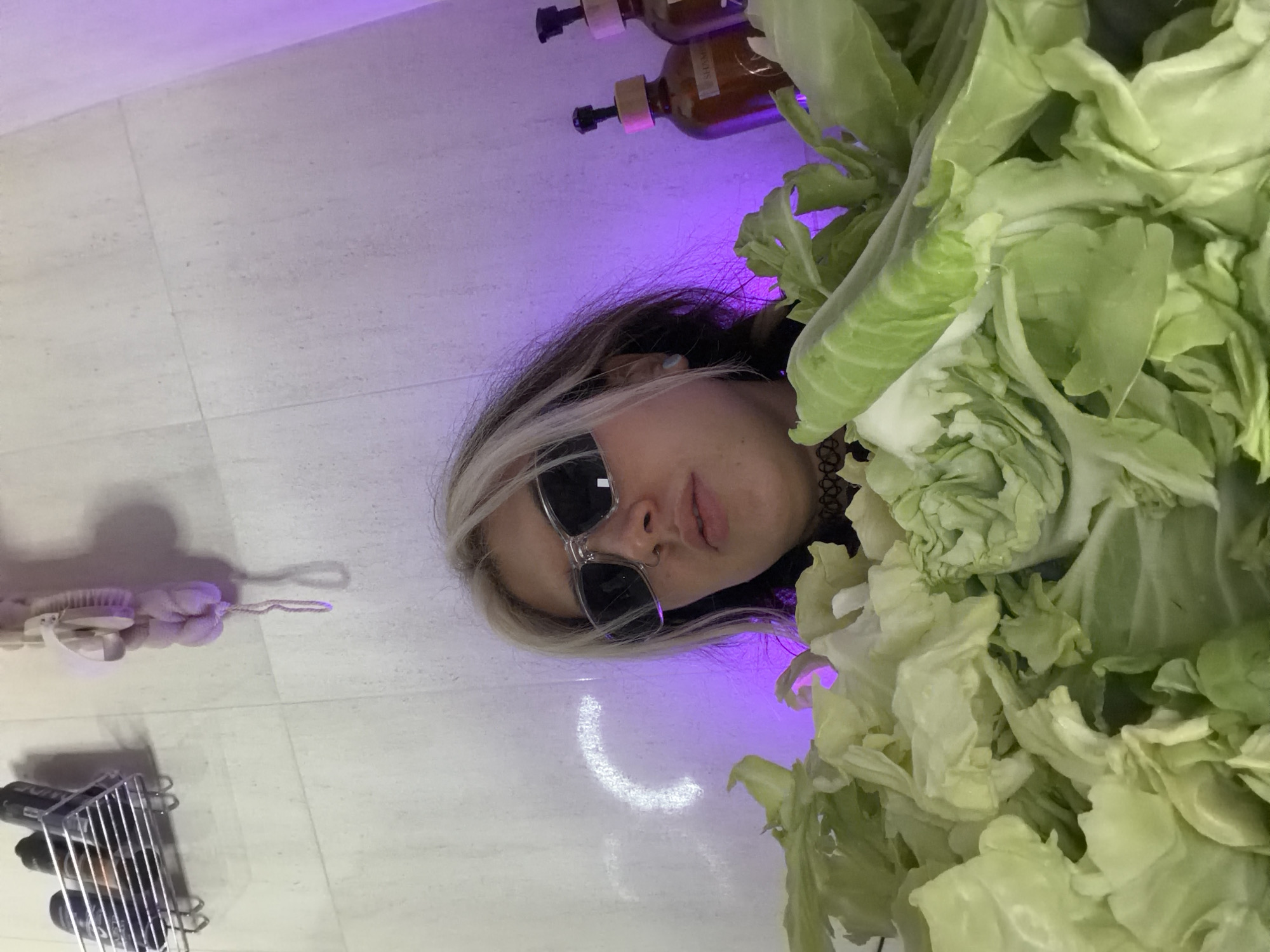 An individual with dark hair and blonde streaks is depicted in an indoor setting, submerged up to the chest in a large quantity of light green cabbage leaves. The person wears clear-framed, dark-lensed sunglasses and a dark patterned choker, looking directly forward with a neutral expression. A light blue earring is visible in the left ear. The background consists of white tiled walls. On the upper left, a chrome shower caddy holds several bottles. To its right, a pink bath pouf hangs. On the upper right, two amber-colored pump bottles are visible, illuminated by a purple light source that casts a soft glow across the right side of the scene. The visual information within the image does not provide specific details for identifying a precise location within the city of Budva, Montenegro.