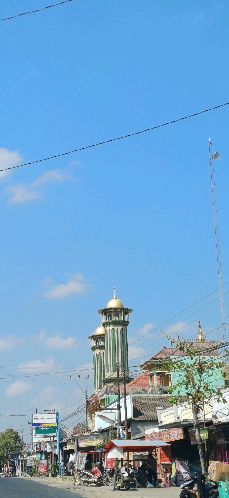 This image captures a street scene in Pucakwangi, Indonesia, characterized by a clear blue sky with sparse clouds. Dominating the upper middle portion of the frame are two distinct minarets of a mosque, featuring green and white stripes and topped with golden domes. To the right, a tall transmission tower with a satellite dish is visible. The foreground shows a street lined with various buildings and shops. Several motorcycles are parked along the roadside, and a few individuals are present, appearing to be engaged in activities related to the shops or street vendors. Power lines crisscross the scene, indicating the presence of electrical infrastructure. The overall environment suggests a typical urban or semi-urban setting in Indonesia.