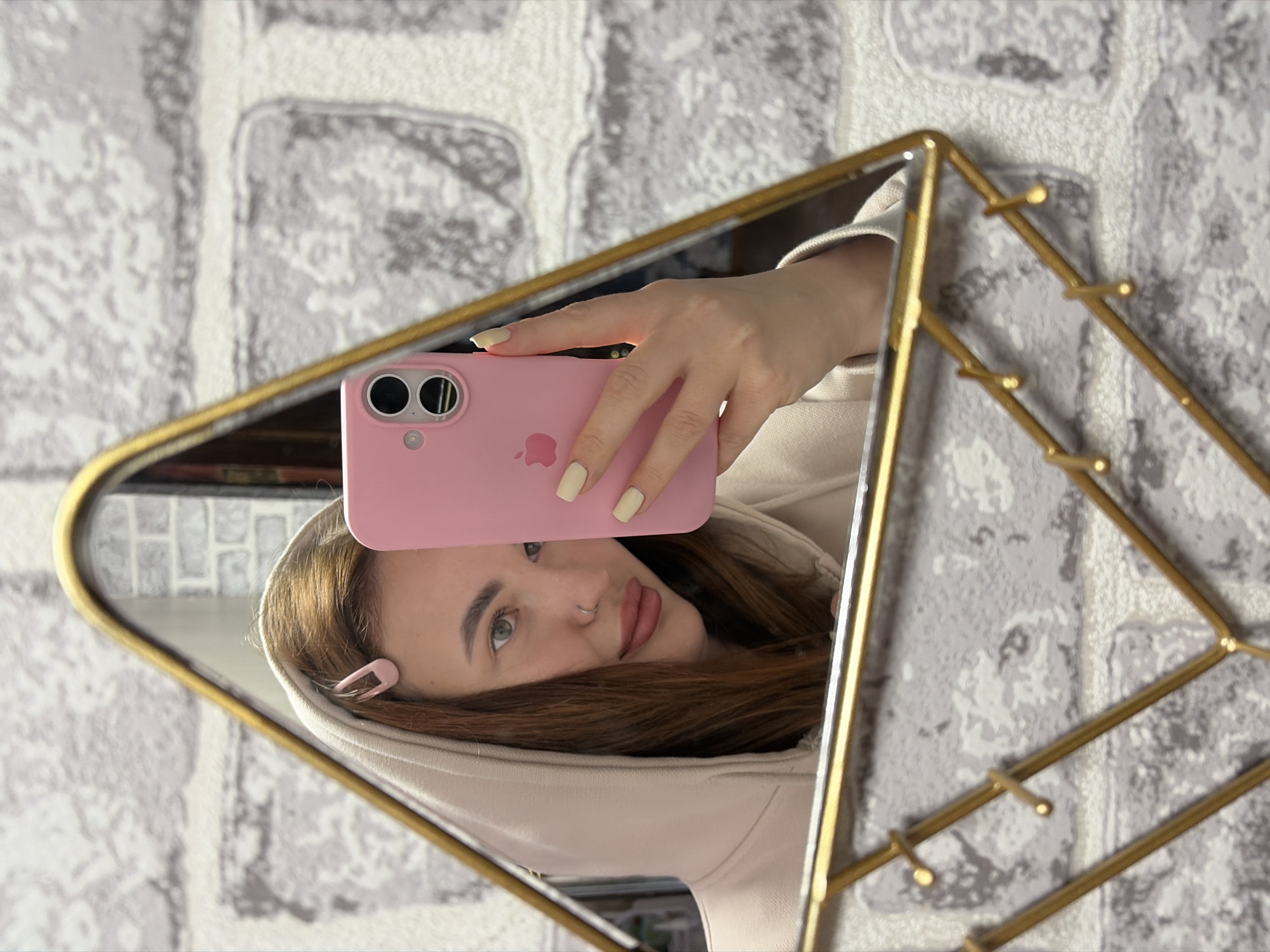 A person is taking a selfie in a mirror. The person is wearing a light-colored hoodie and holding a pink iPhone. A pink barrette is visible in their hair. The mirror has a gold-colored frame and is reflecting a grey brick-patterned wall. The location is Novopokrovskaya, Russia.