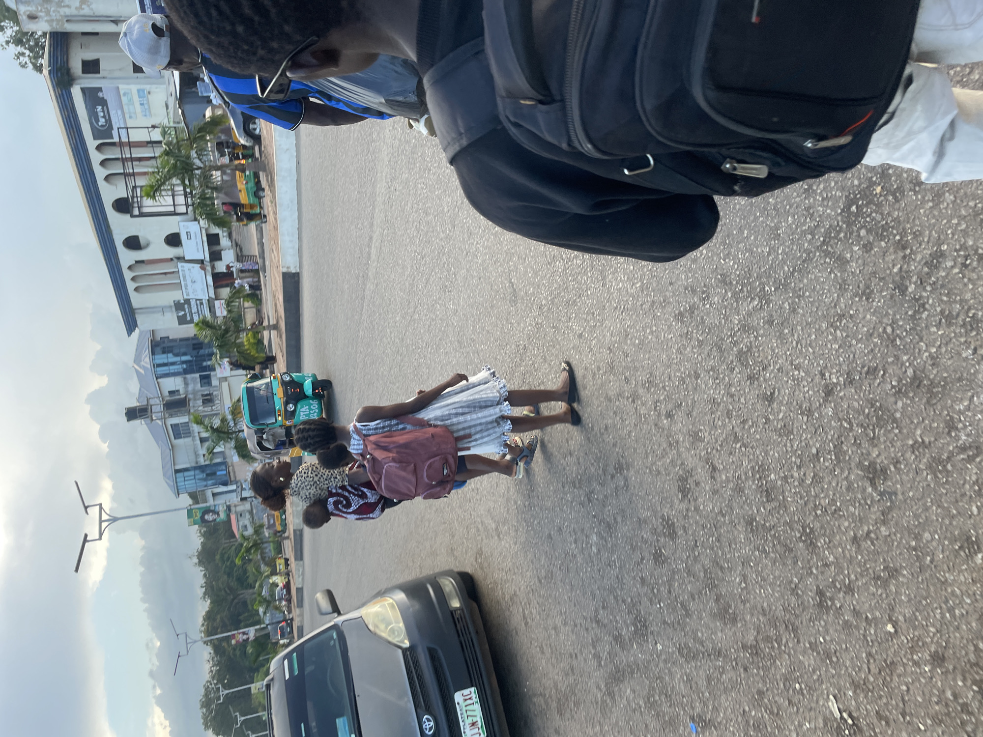 A medium shot captures a street scene in Barakin Lerawi, Nigeria. The foreground is dominated by the back of a person wearing a black sweatshirt and carrying a black backpack, with light-colored pants visible. To the left, a dark-colored sedan is parked. Further into the scene, a woman is walking, carrying a baby on her back. A young girl, wearing a white and gray striped dress and a maroon backpack, walks beside her. A teal-colored auto-rickshaw, a common mode of transport in Nigeria, is visible on the road. In the background, buildings with modern architecture line the street, and palm trees are scattered throughout the environment. The sky is overcast with clouds, suggesting it is either early morning or late afternoon.