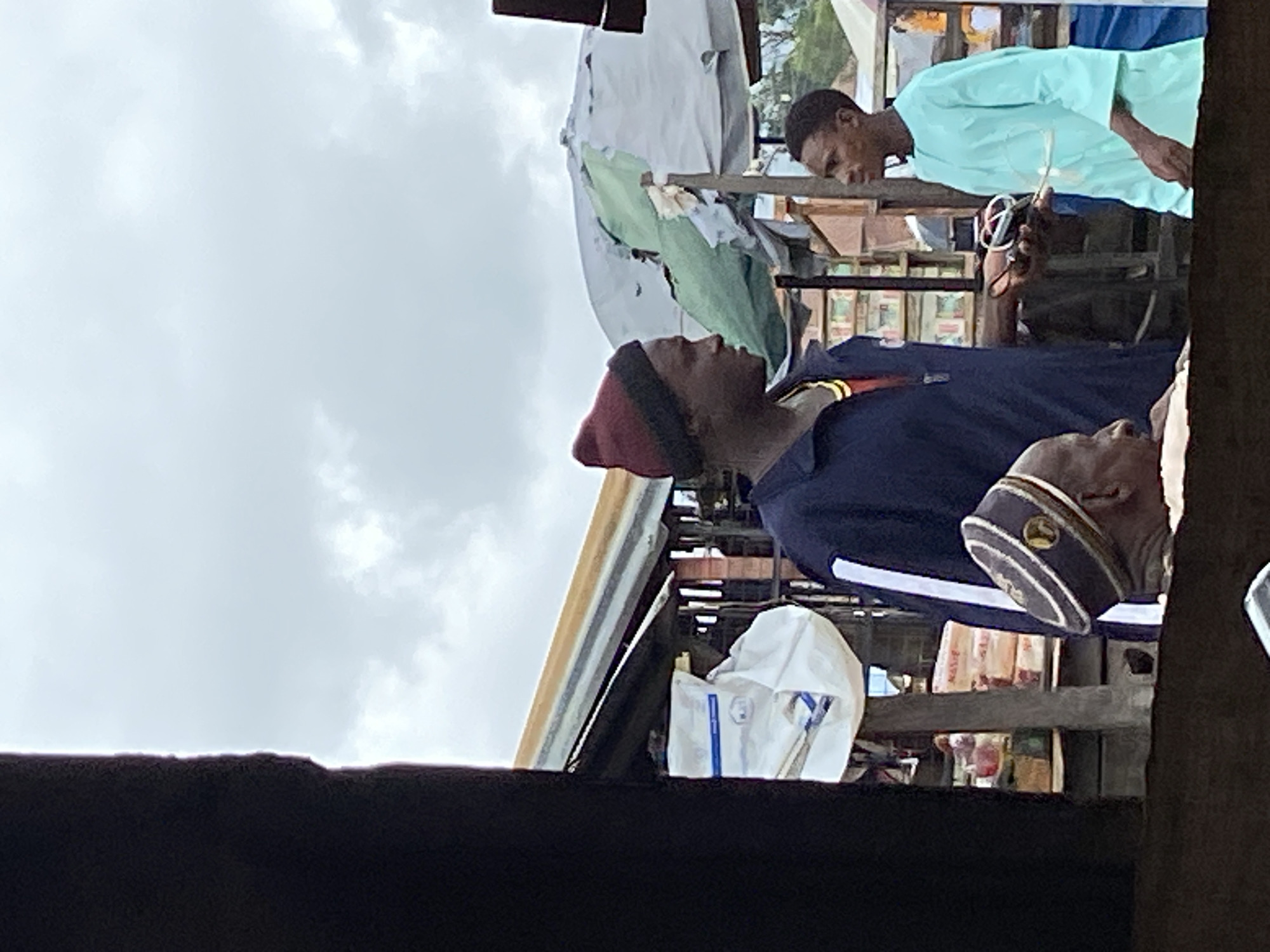 In Mararaba, Nigeria, the image depicts three African men outdoors. One man wears a red cap and a blue shirt with a white stripe, another a light blue top, and the third, partially visible, wears a dark cap with a decorative band. Laundry hangs overhead. The scene appears to be in a market or informal commercial area.