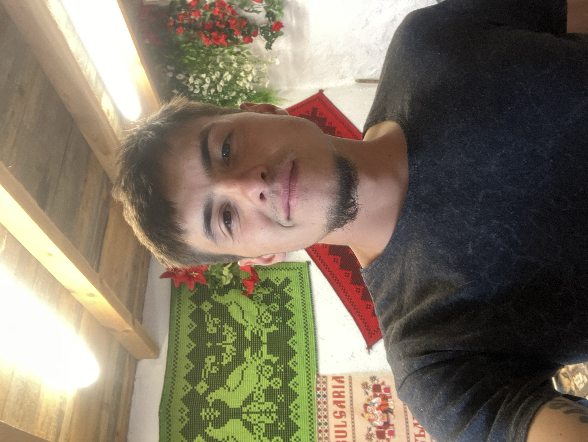 A man is posing indoors in what appears to be a traditional Bulgarian setting. The background includes various textiles, including one with "BULGARIA" printed on it, and another green woven textile. Flowers and foliage decorate the wall, suggesting a celebratory or decorative atmosphere. The man is wearing a dark grey t-shirt. The location is likely in or near Komuniga, Bulgaria, based on the traditional Bulgarian elements and the text on one of the textiles. The lighting is somewhat uneven, with bright light sources in the upper left corner of the image.