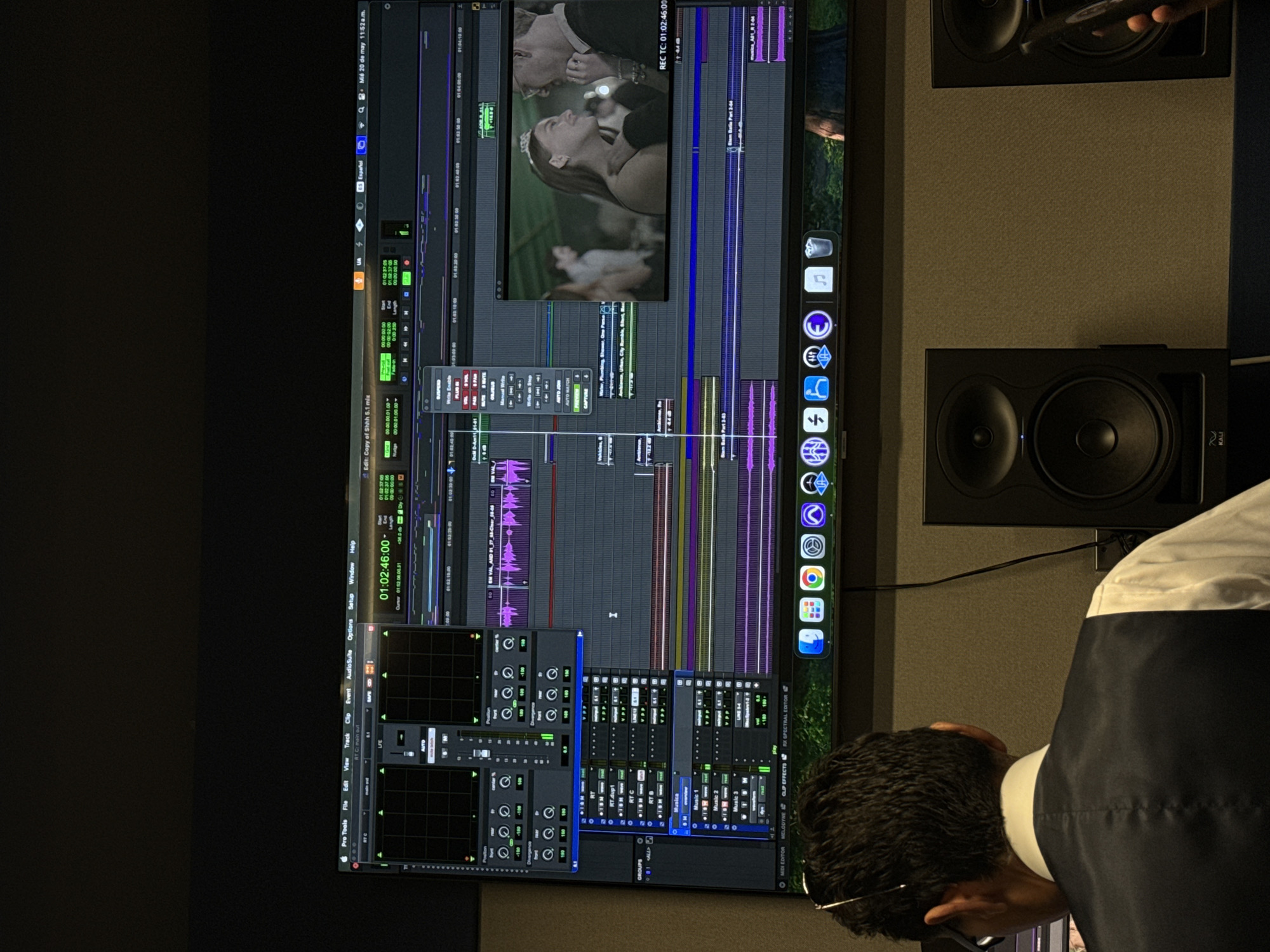 The image captures a person, likely a sound engineer or video editor, working in a professional audio or video post-production environment. The person, dressed in a shirt with a vest or jacket, is facing a large computer monitor that displays the editing interface of Pro Tools, a digital audio workstation software. The Pro Tools interface shows a timeline, audio waveforms, video preview (possibly of a wedding), and various mixing and editing tools.
The room appears to be a sound-treated studio, indicated by the two professional-grade studio monitors mounted on the wall. The wall behind the monitor is covered in sound-absorbing material, suggesting that this space is designed for critical listening and audio editing.
The location is likely a post-production studio, but without further clues, the city cannot be determined.