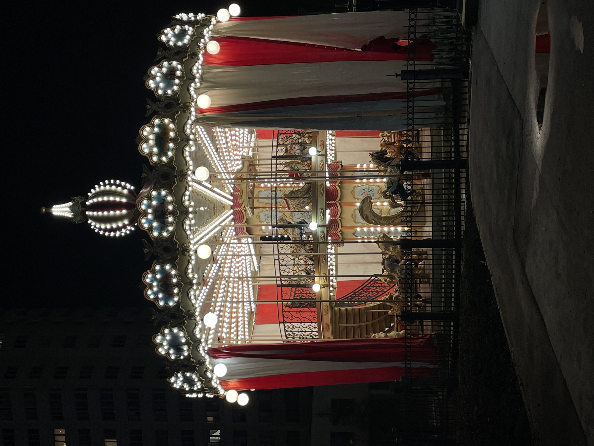 At night, a carousel stands illuminated by numerous lights, with a red, white, and silver canopy at its top. The carousel is enclosed by a wrought-iron fence, and part of a modern building is visible in the lower left corner of the frame. To the right of the carousel is a concrete walkway, while the sky is black. The carousel is located in Moscow, Russia.