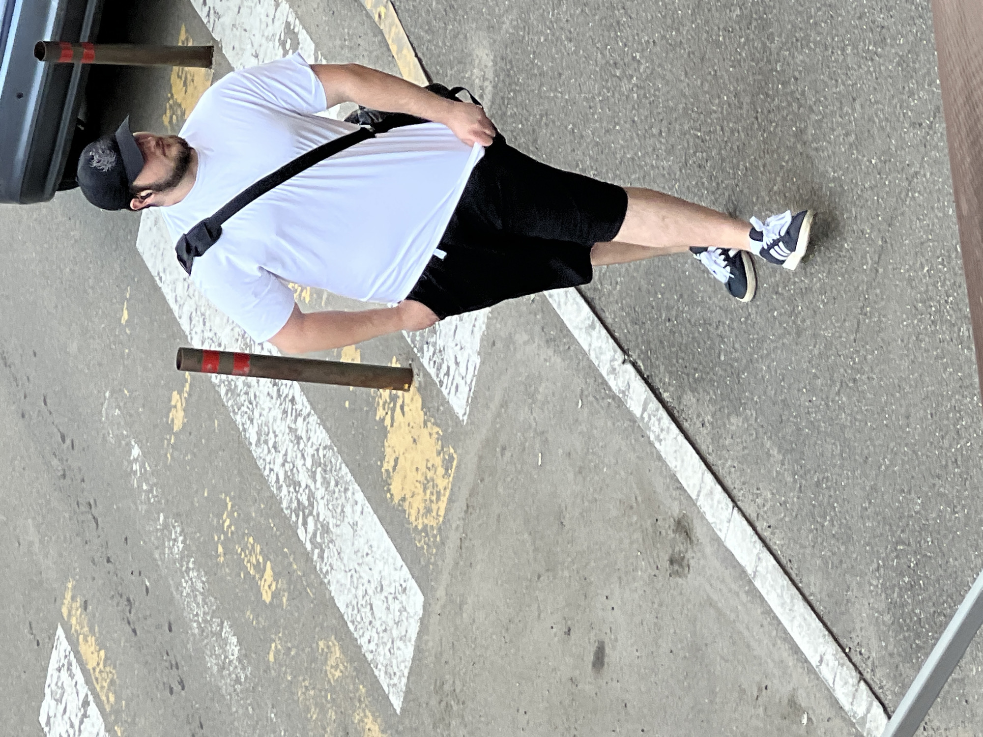 A man is captured from an elevated perspective, standing on what appears to be an asphalt street or parking lot. He is dressed in a casual outfit consisting of a plain white short-sleeved t-shirt, black shorts, and black sneakers with white laces and distinct white stripes on the side. He also wears a black baseball cap with a subtle white or grey design on the front and has a beard. A black cross-body bag is slung over his shoulder. His right hand is near his shorts, possibly adjusting them or his t-shirt.
The ground features prominent white and faded yellow road markings, suggesting a zebra crossing or parking area. To the left, a dark grey car bumper is partially visible. Two metal bollards, each with a red band near the top, are seen; one is behind the car, and another is further down to the left of the man. A sharp white line runs along the right edge of the frame, possibly delineating a curb or boundary. The lighting suggests it is daytime, with no harsh shadows indicating strong direct sunlight. No discernible text is visible on the man's clothing or in the surroundings. The man's expression is not visible as he is looking away from the camera. The setting is consistent with an urban or suburban outdoor environment in Krasnodar, Russia.