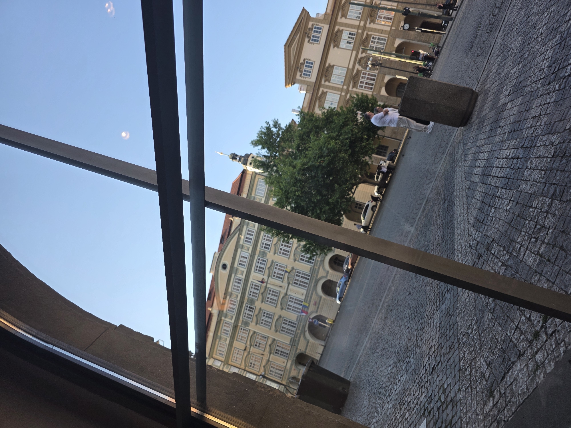 The image shows a view of a city street through a window. The street is paved with cobblestones. Buildings with multiple windows and architectural details line the street. A green tree is visible in front of one of the buildings. A man in white clothes stands near a concrete post on the street. Several cars are parked along the street. The sky is blue. The location is in an unknown city.