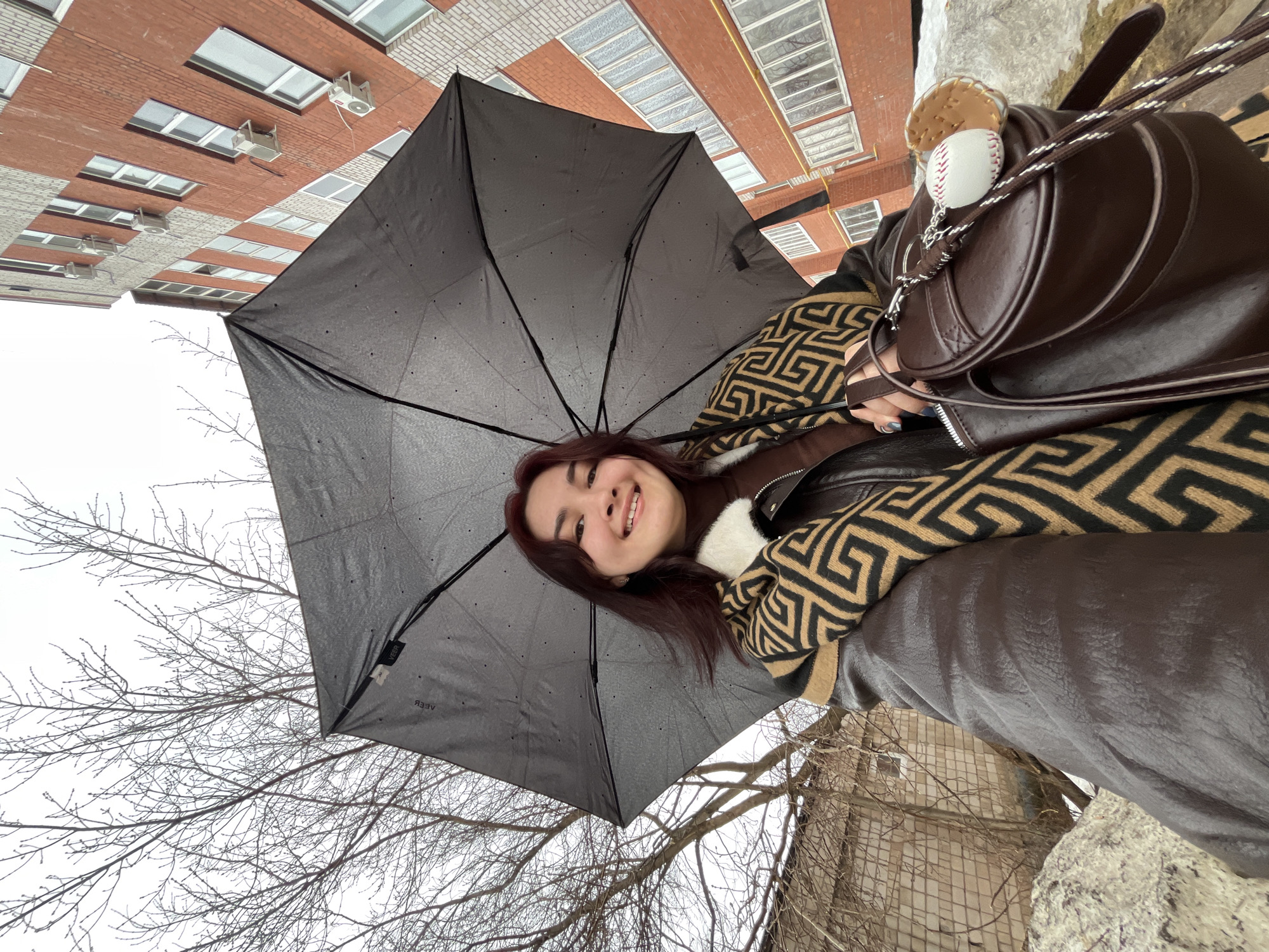A young adult female is depicted in a low-angle outdoor selfie, smiling towards the camera. She has dark reddish-brown hair and is wearing a brown turtleneck, a dark brown leather-look jacket with a white fur collar, and a patterned scarf featuring a black and tan geometric design. Her left hand holds a black umbrella, partially open, with the brand "VEER" visible on its underside. Her right hand supports a dark brown, rounded bag, which has a keychain consisting of a miniature white baseball with red stitching and a miniature tan baseball glove.
The background on the left features a bare deciduous tree with numerous branches against an overcast sky. On the right, a multi-story building constructed of red-orange and lighter-colored bricks is visible, featuring multiple windows and several external air conditioning units affixed to its facade. The ground in the foreground and lower left appears to be covered with an irregular, light-colored substance, consistent with snow or debris. A brick wall or fence is partially visible behind the tree. The scene takes place in Neftekamsk, Russia.