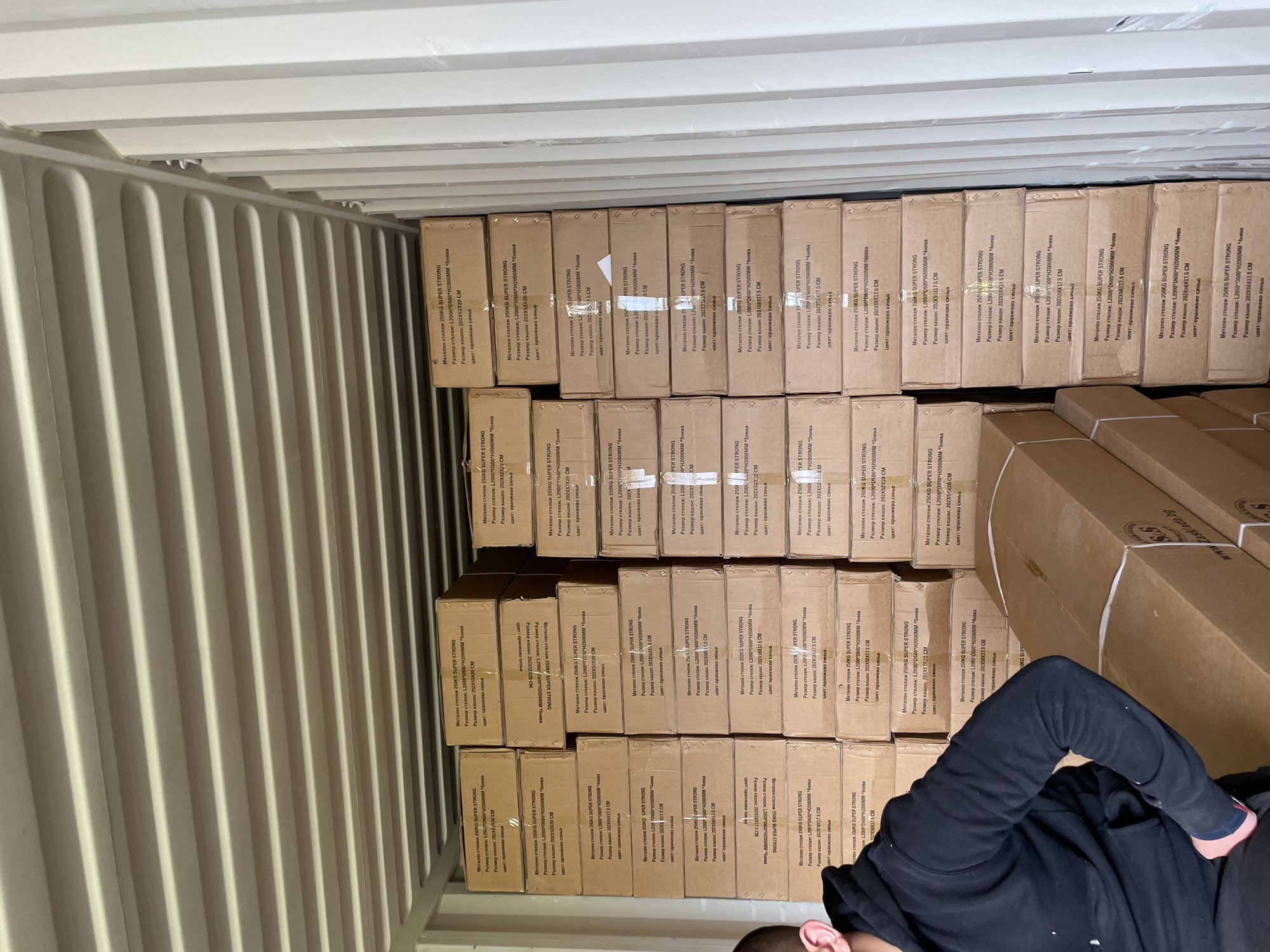 The interior of a shipping container is packed with cardboard boxes. The boxes are stacked tightly against one wall. The boxes have text printed on them, including "Метален стелаж 250KG SUPER STRONG". The text also includes dimensions in millimeters and centimeters, as well as the phrase "цвят: оранжево синьо". A person in a black hooded sweatshirt is visible at the bottom right of the frame, near the stack of boxes. The walls of the container are corrugated metal. The top of the container is also visible, showing the ceiling structure. There is no information in the image to confirm the location of the city of Asenovgrad, Bulgaria.