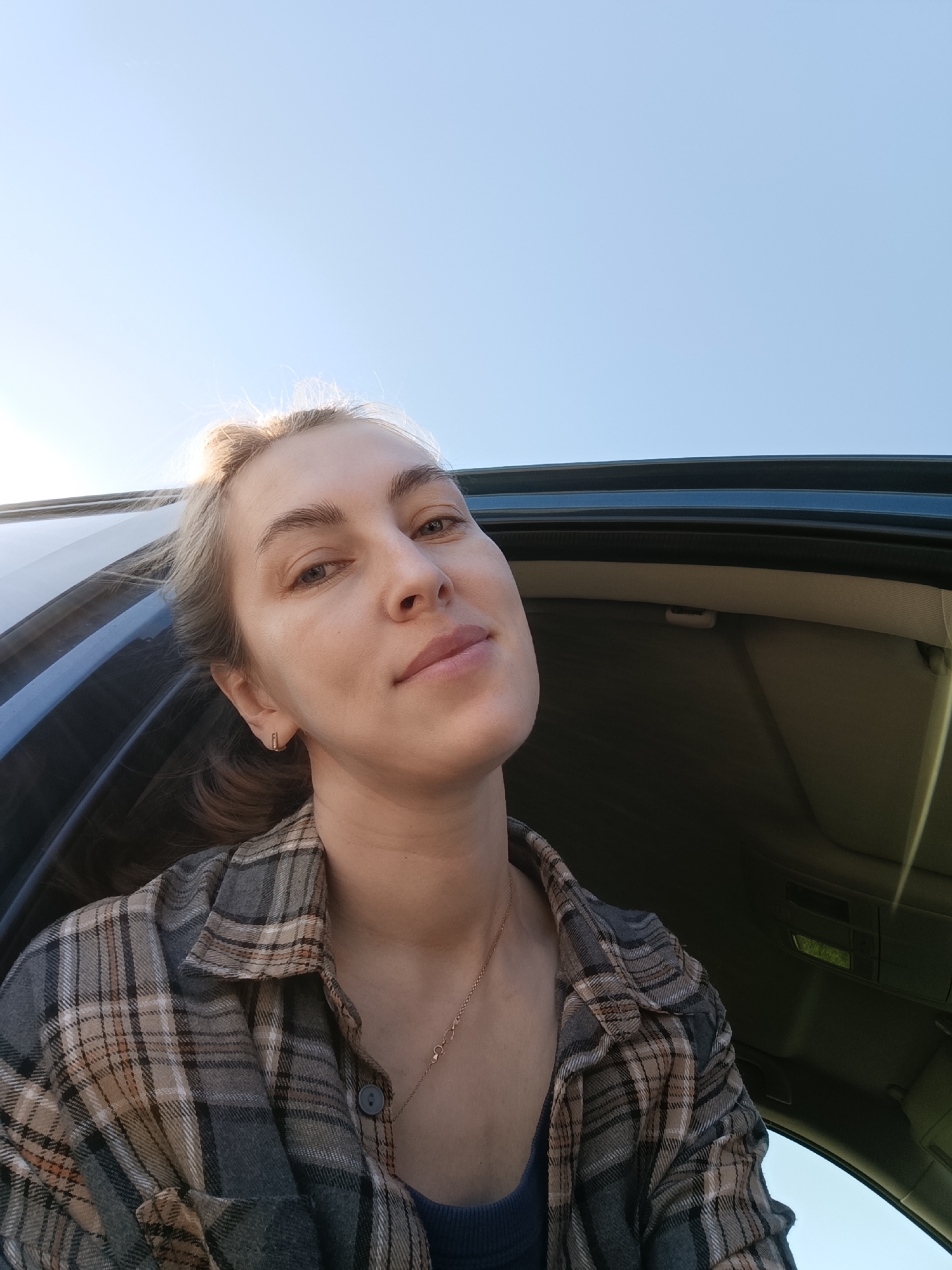 A medium shot depicts a young woman inside a car, appearing to take a selfie. The woman has fair skin, light blonde hair, and is wearing a plaid flannel shirt over a blue top, along with a delicate gold necklace. She has a small rectangular earring in her left ear. The interior of the car is visible, with a dark-colored trim and lighter fabric on the ceiling. Outside the car, a clear blue sky is visible. The location is assumed to be Chaikovsky, Russia.