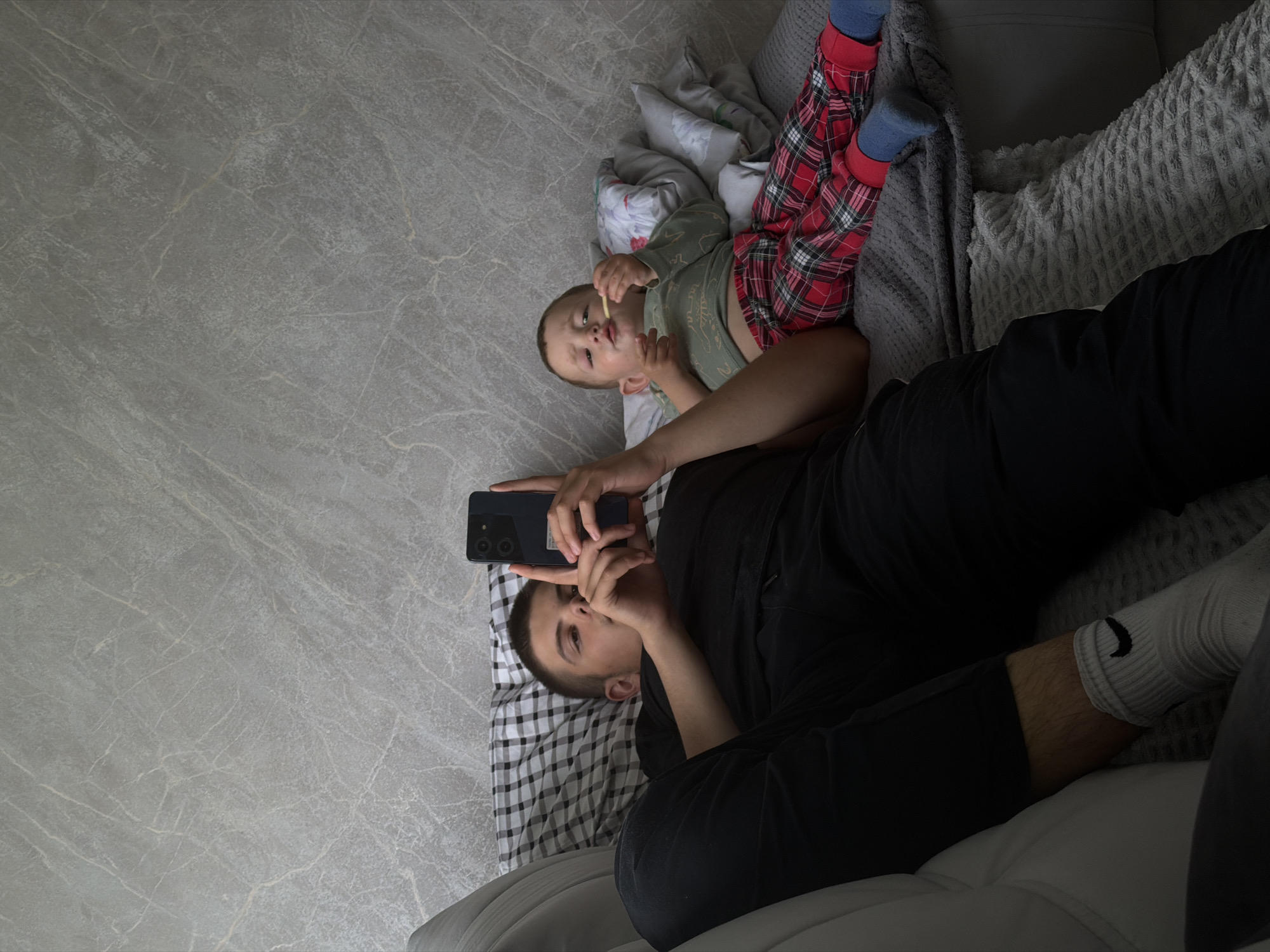 Two individuals are present in a domestic setting, lying on a sofa. The person on the left, a male, is facing forward and holding a smartphone. He is wearing a black t-shirt, black pants, and white socks with a black Nike logo. The person on the right, a child, is lying on their side and eating. The child is wearing a light green long-sleeved shirt and red and black plaid pants with blue socks. The background is a textured gray wall. The scene appears to be indoors.