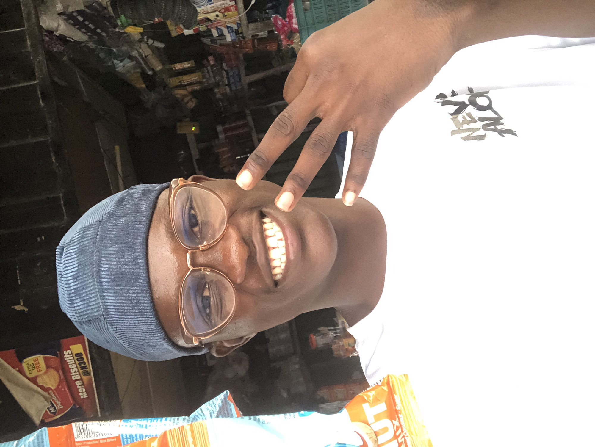 A medium shot captures a young Black male wearing a blue beanie and round, rose-gold rimmed glasses, smiling broadly at the camera. He is wearing a white t-shirt with a graphic design of the words "NEV ALO" in black brush strokes on the left side of his chest. His right hand is raised, with his index, middle, and ring fingers extended in a peace sign. The background appears to be an outdoor stall or market in Ado, Nigeria, with various packaged goods visible on shelves. To the left of the subject, a partially visible orange and white package of biscuits can be seen. The lighting suggests it is daytime.