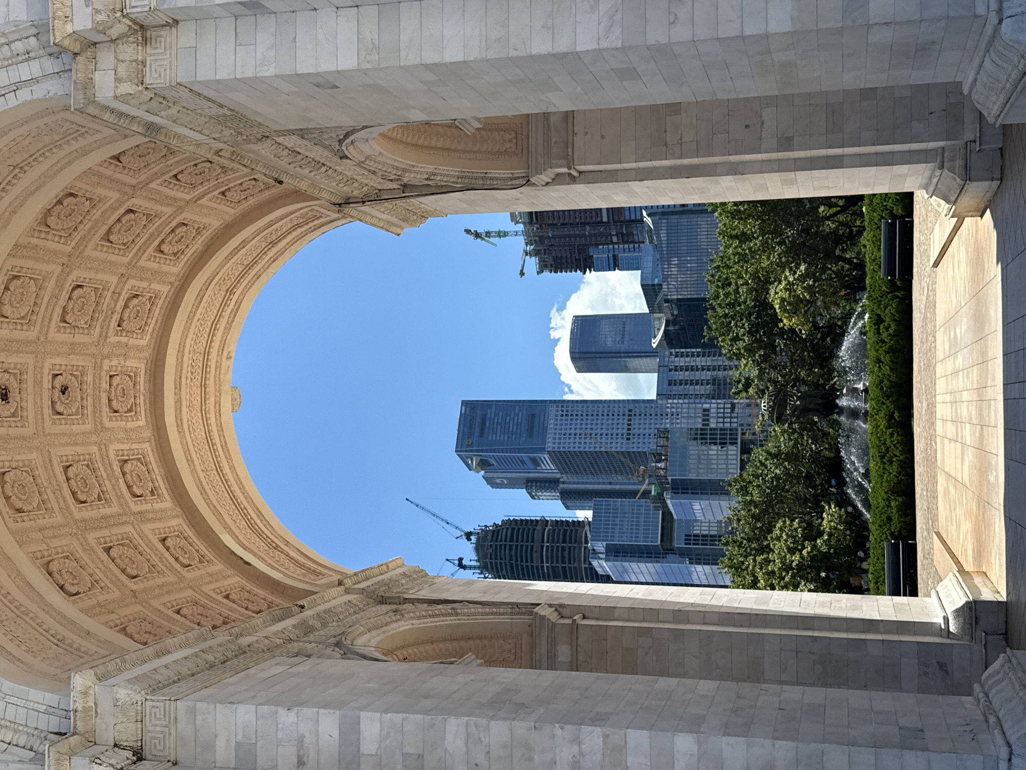 This image, taken in Nanshan District, China, captures a view through a grand marble archway towards a modern cityscape. The archway's interior is ornately decorated with a coffered ceiling featuring floral motifs. The exterior of the arch is constructed from large, pale stone blocks, with classical architectural details visible along the top and sides.
Beyond the arch, a bright blue sky with scattered white clouds serves as the backdrop for a cluster of contemporary skyscrapers. One prominent building, identified by "SHENZHEN" signage, is tall and slender with a dark glass facade. Adjacent to it, other buildings exhibit varied designs, with some featuring visible construction cranes, indicating ongoing development. A circular fountain sprays water in the middle ground, surrounded by lush green bushes and trees. Two dark park benches are positioned on a paved walkway in front of the greenery. The overall scene suggests a blend of historical architecture and modern urban development within a vibrant city. There are no people visible in the image.