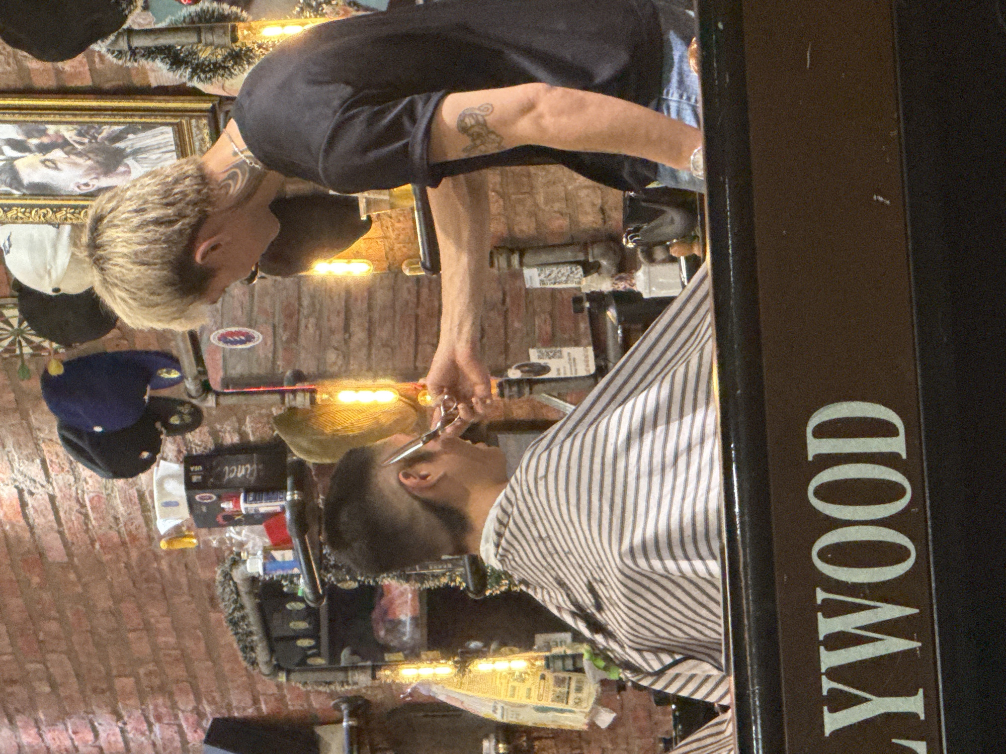 In Đà Nẵng, Vietnam, the image shows a male barber with blonde hair and tattoos cutting a customer's hair in a barbershop. The barber wears a black t-shirt and jeans. The customer is draped in a striped barber cape. Tools and products are visible on shelves along a brick wall. A sign with "LYWOOD" is partially visible. Hats and a framed picture are among the decorations.