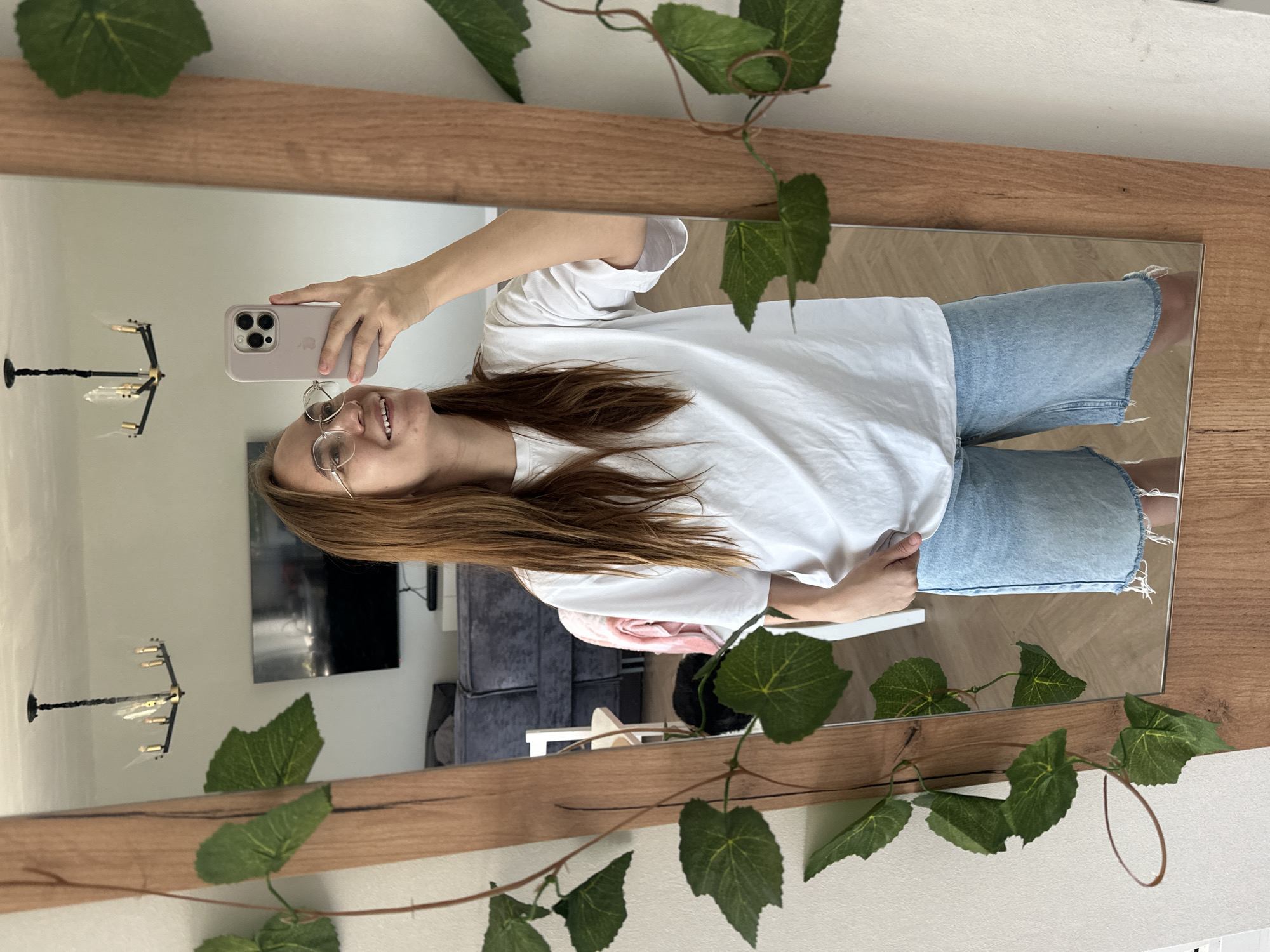 A woman is taking a mirror selfie. She has long, light brown hair and is wearing glasses, a white t-shirt, and light blue denim shorts. She is holding a phone with a light-colored case. The mirror has a wooden frame and is decorated with faux green vines. The reflection shows a room with a television, a couch, and a chandelier. The location cannot be determined to be Kharnavy, Russia from the image provided.